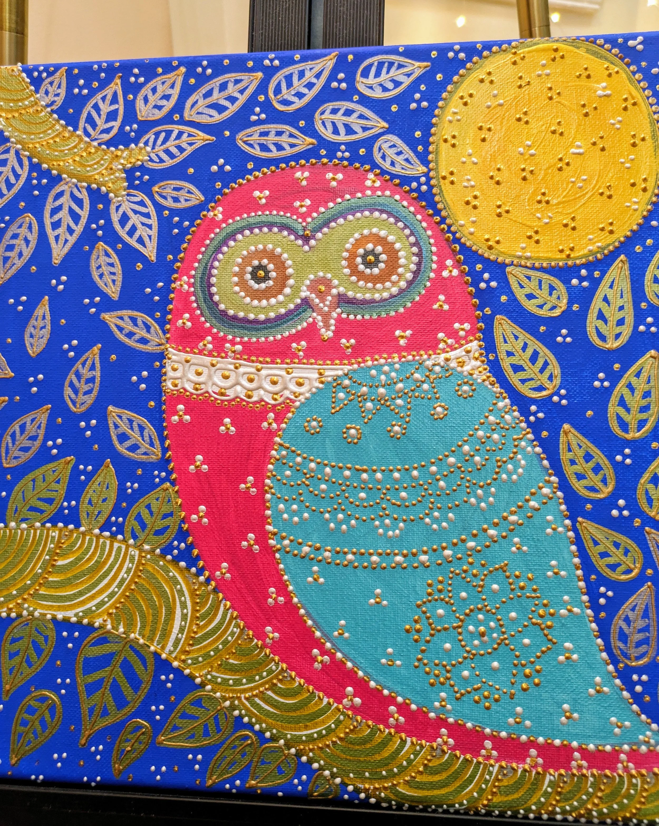 Decorative artwork of an owl with pink and teal body, gold and white accents, surrounded by gold leaves, a gold crescent, and a gold full moon on a blue background.
