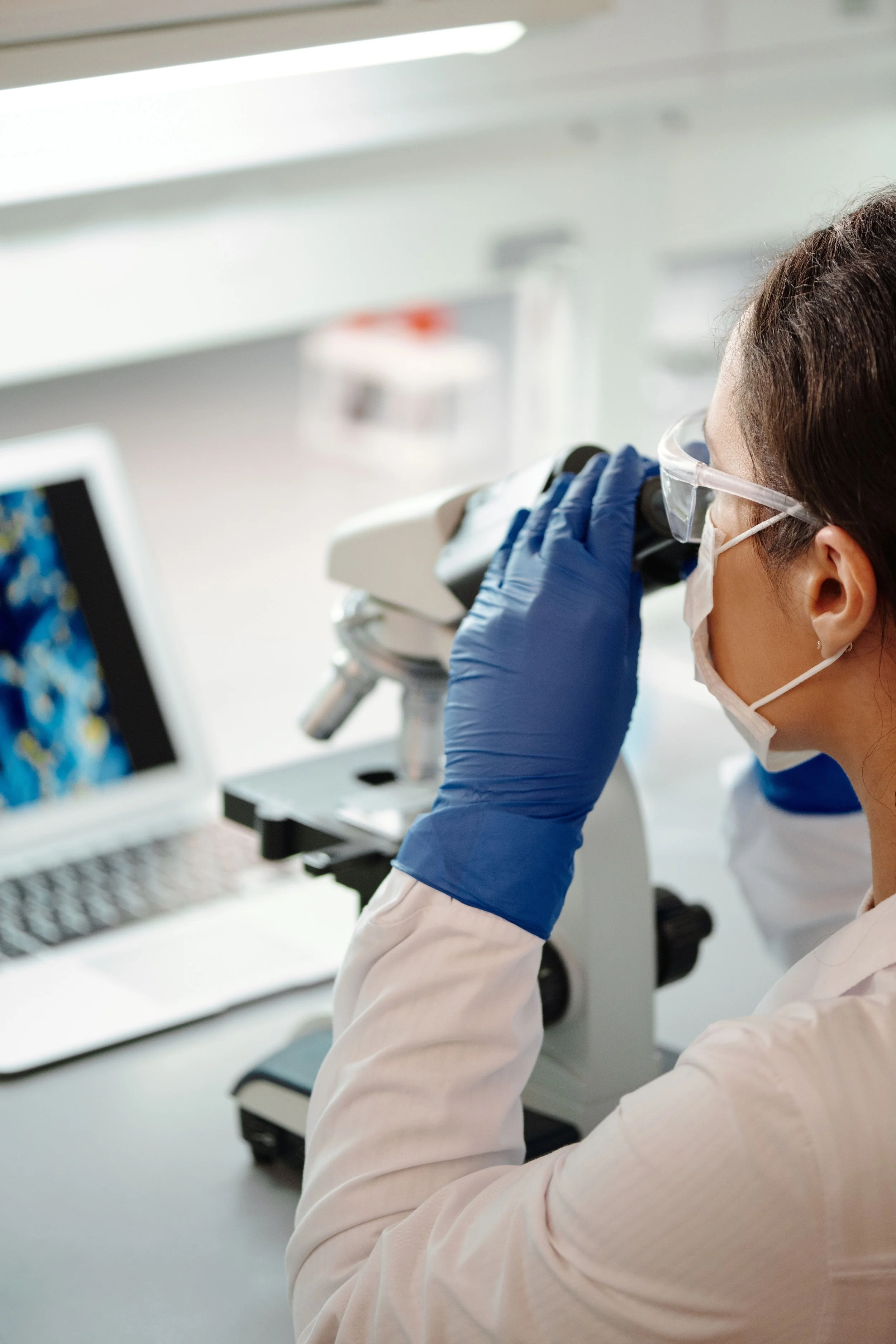 A scientist or researcher looking through a microscope in a laboratory with scientific equipment and a computer monitor in the background.