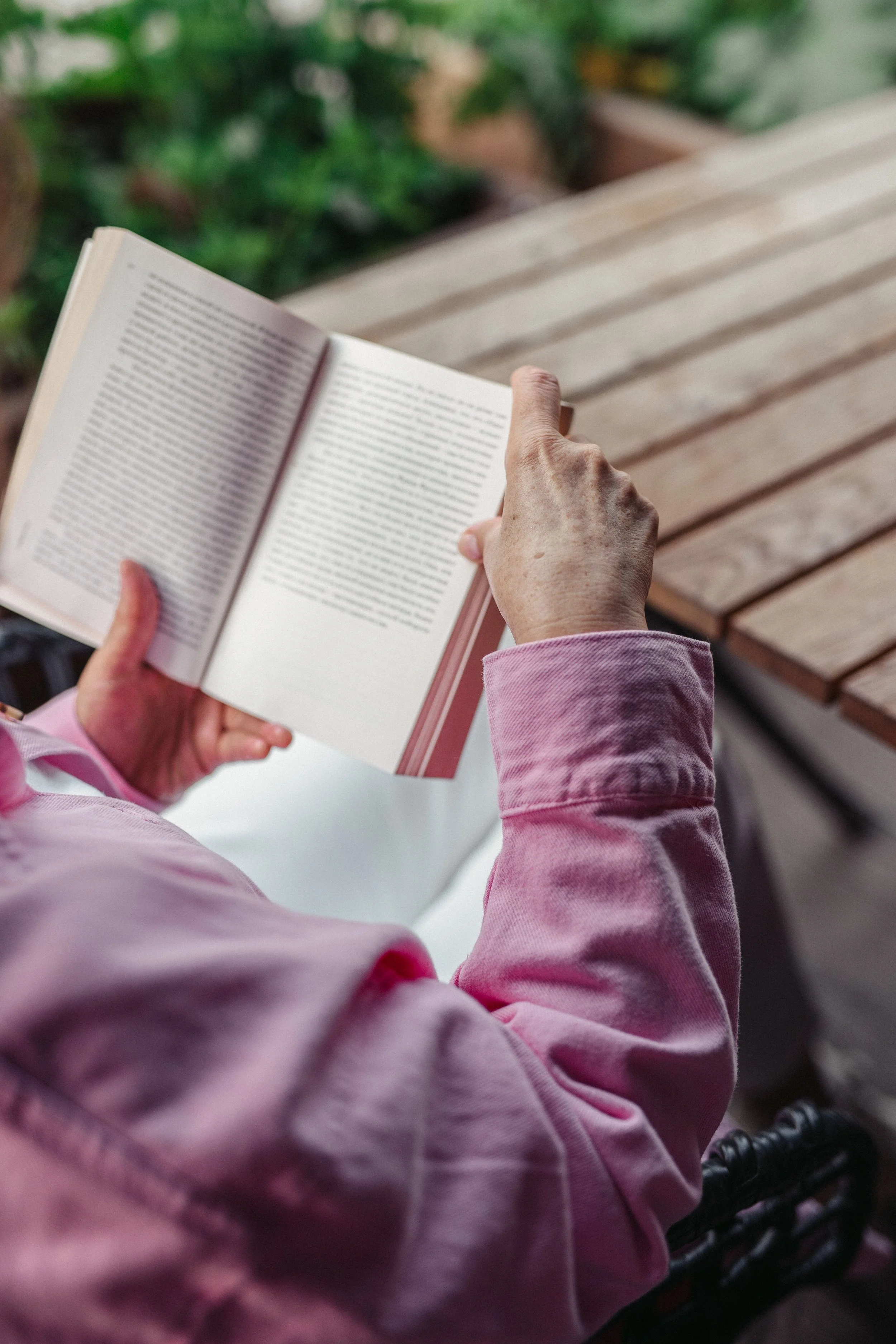 Person wearing a pink long-sleeve shirt or hoodie sitting outdoors, holding an open book with both hands, with a wooden bench and green foliage in the background.