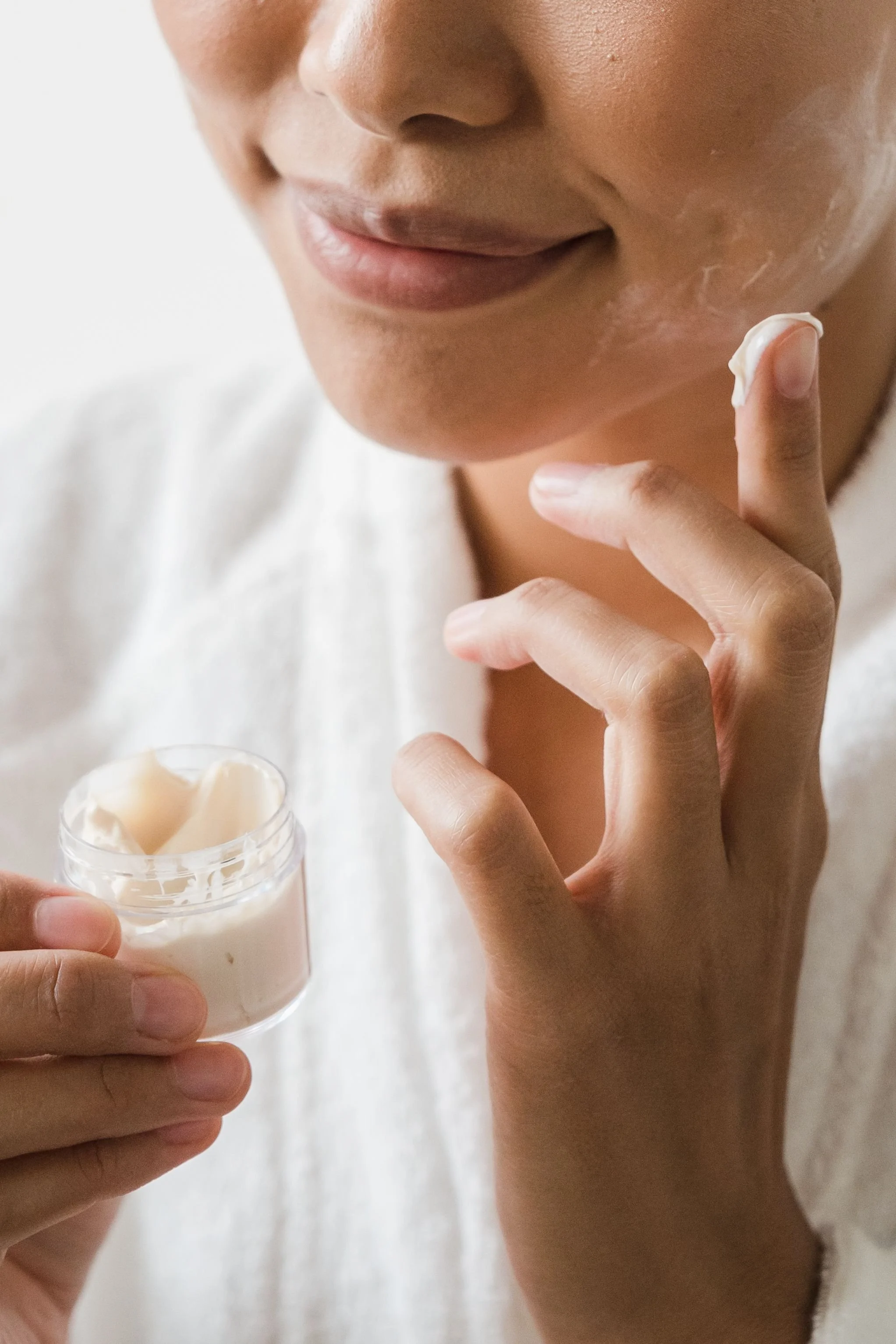 Close-up of a person applying cream to their face with their finger, holding a small container of cream.