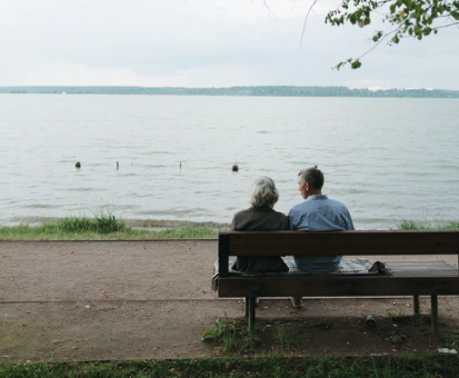 An elderly woman and a middle-aged man sitting on a park bench facing a body of water, with trees and cloudy sky in the background.