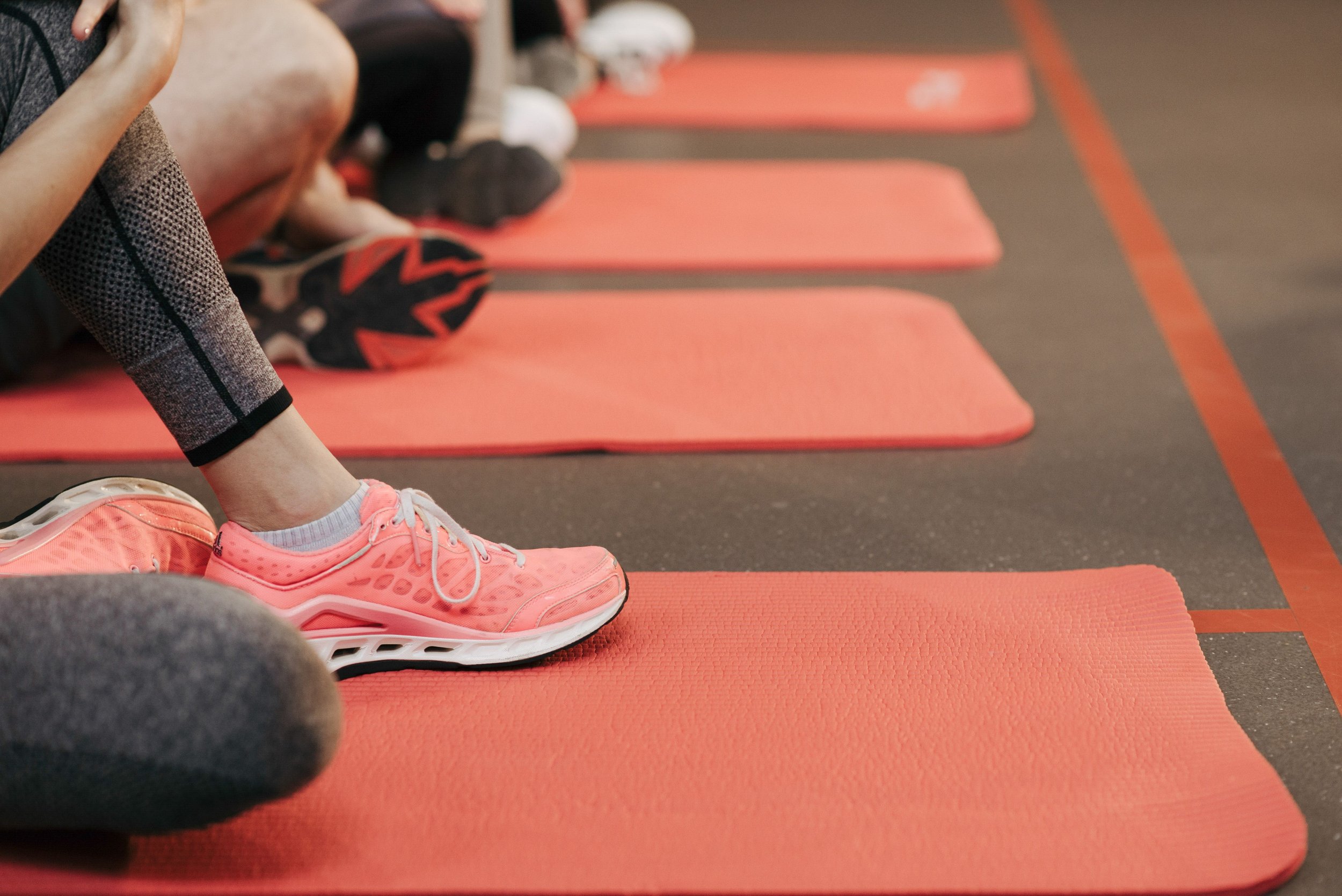 Close-up of a person wearing pink athletic shoes and gray leggings, sitting on an orange exercise mat in a gym with other mats and people in the background.