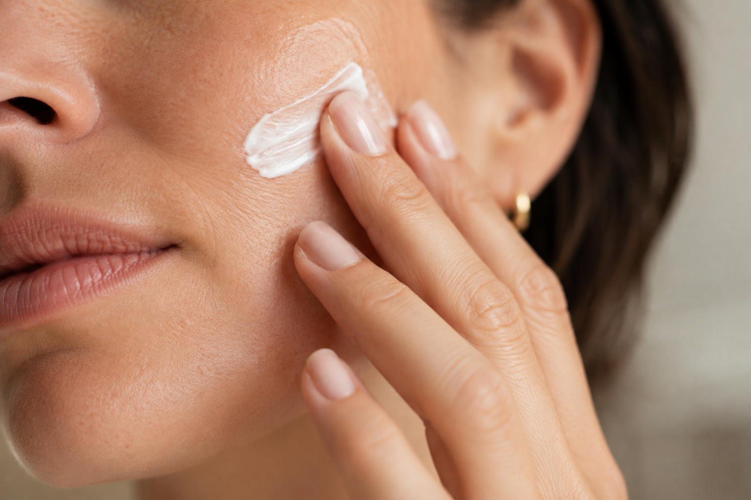A woman applies a white cream or lotion on her face near her cheek with her fingers.