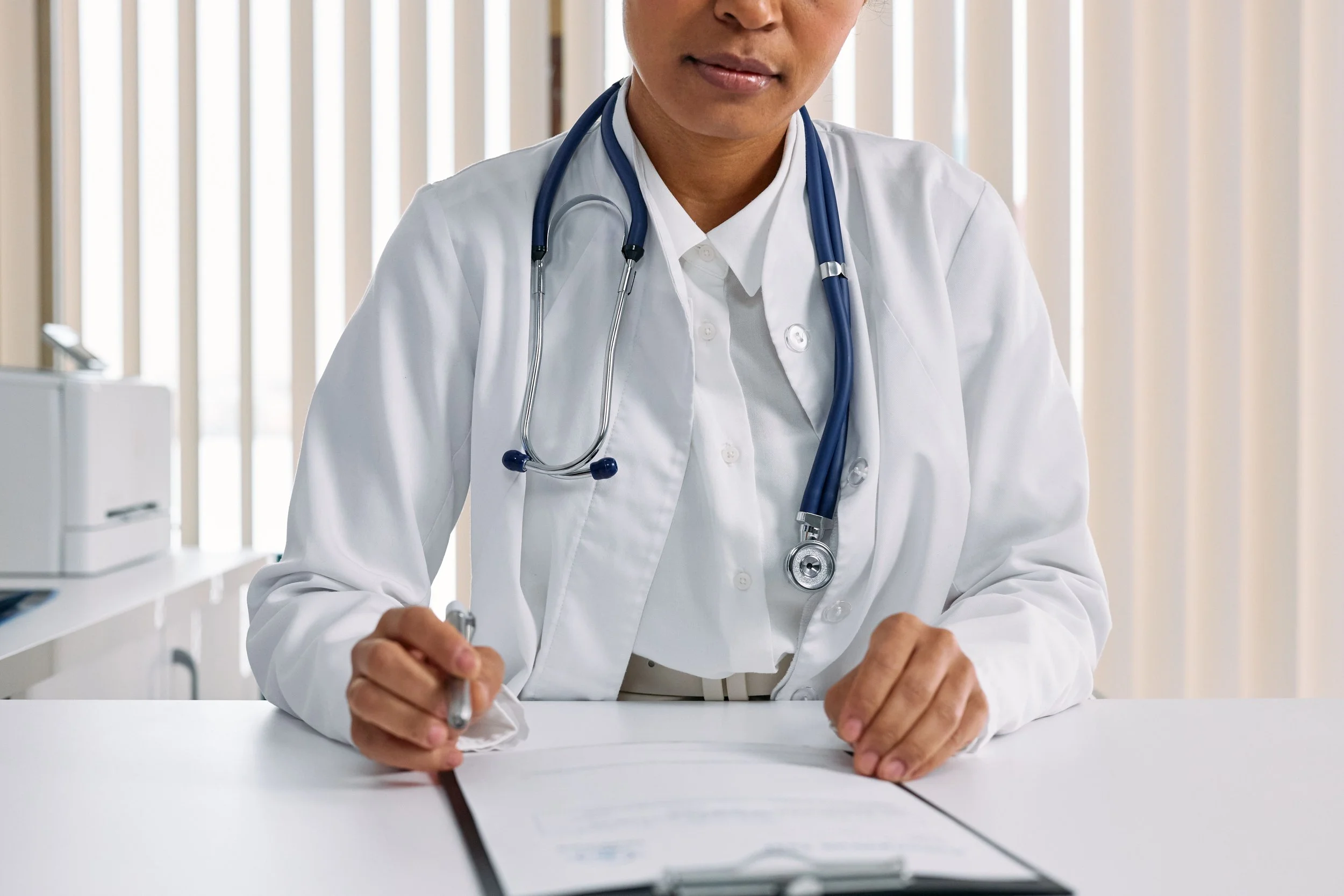 A healthcare professional, wearing a white coat and stethoscope, writing on a piece of paper in a clinical setting.