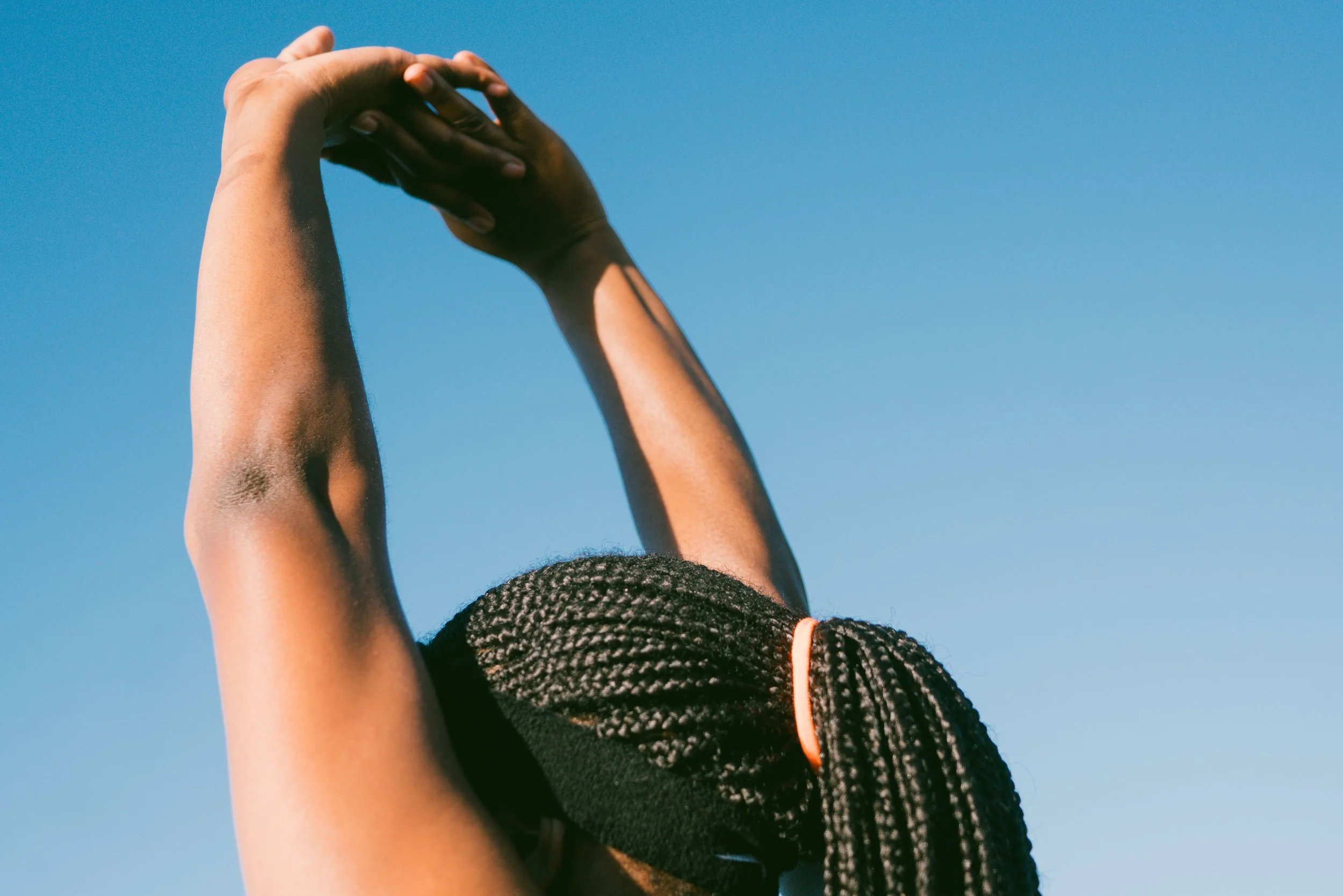 Person with braided hair raising their hands towards a clear blue sky.