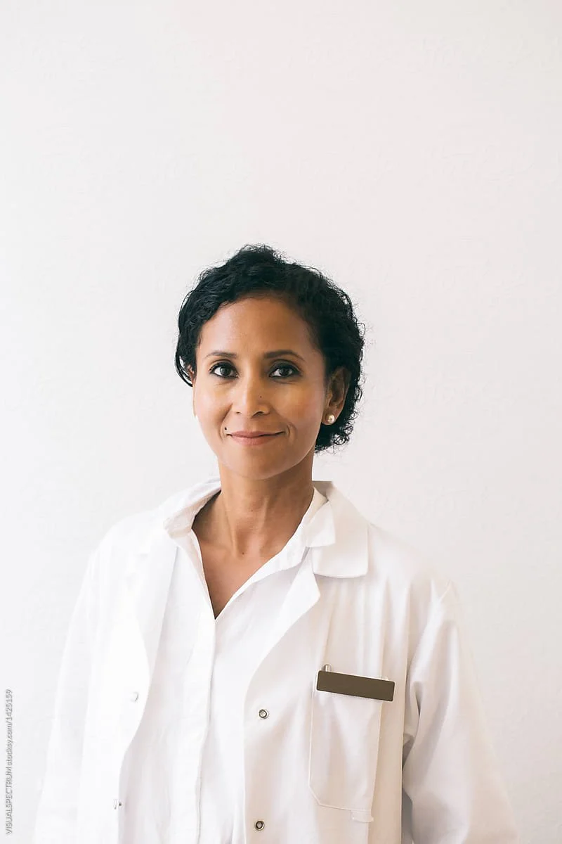 A woman with short curly black hair, wearing a white medical coat, standing against a plain white wall.