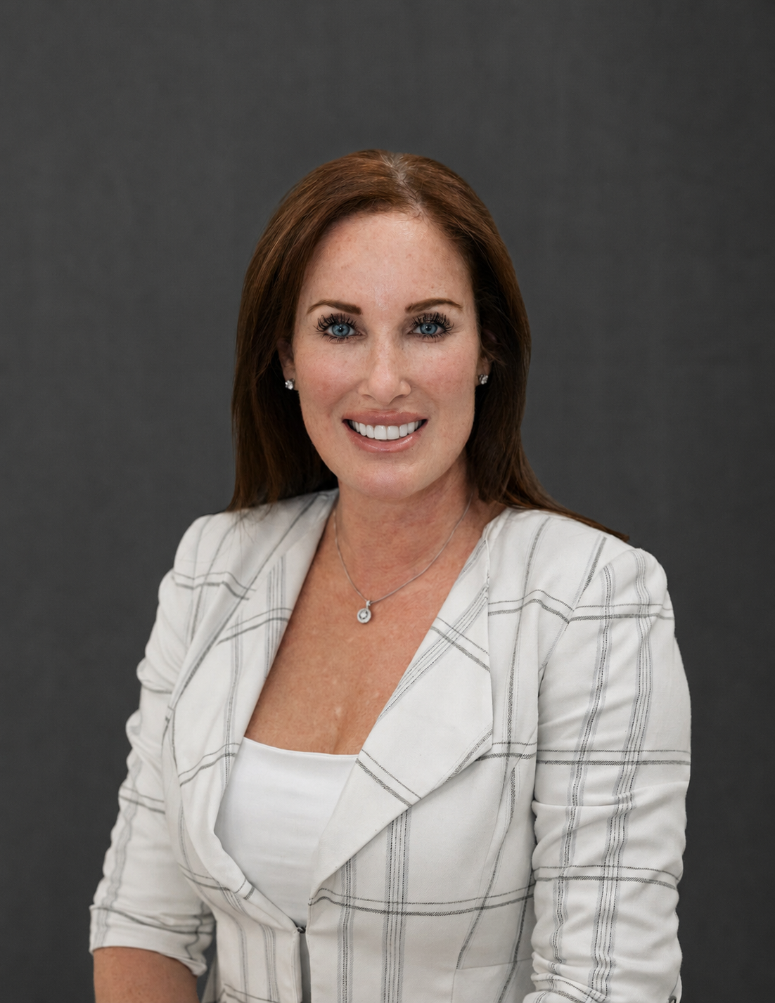 A woman with long red hair, blue eyes, and fair skin, smiling and wearing a white blazer with a checkered pattern over a white top, stands against a dark gray background.