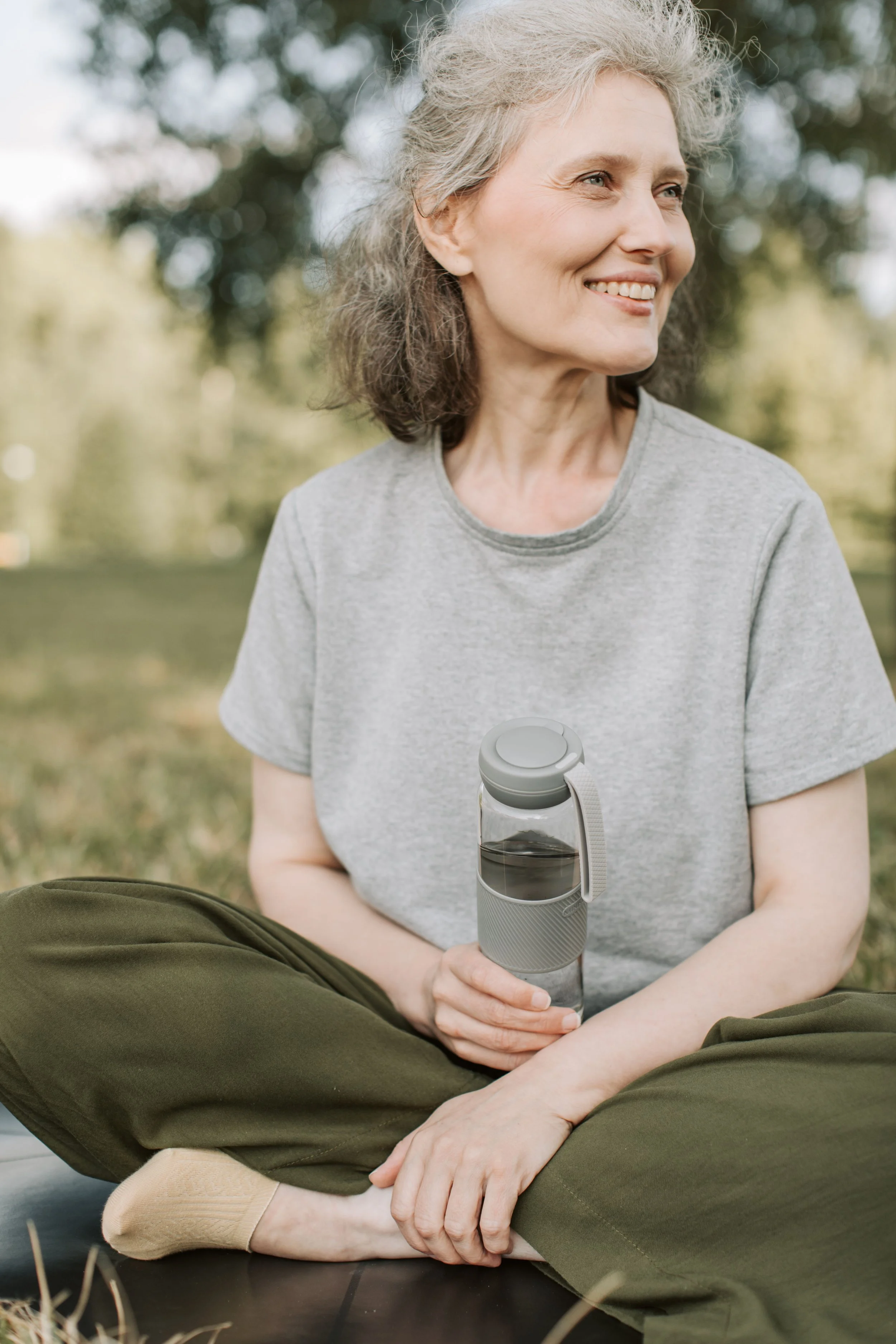 A smiling older woman with gray hair sitting cross-legged outdoors, holding a water bottle, wearing a gray t-shirt and olive green pants.