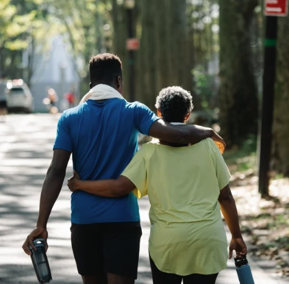 A younger man and an elderly woman walking together in a park, with the man resting his arm on the woman's shoulders.