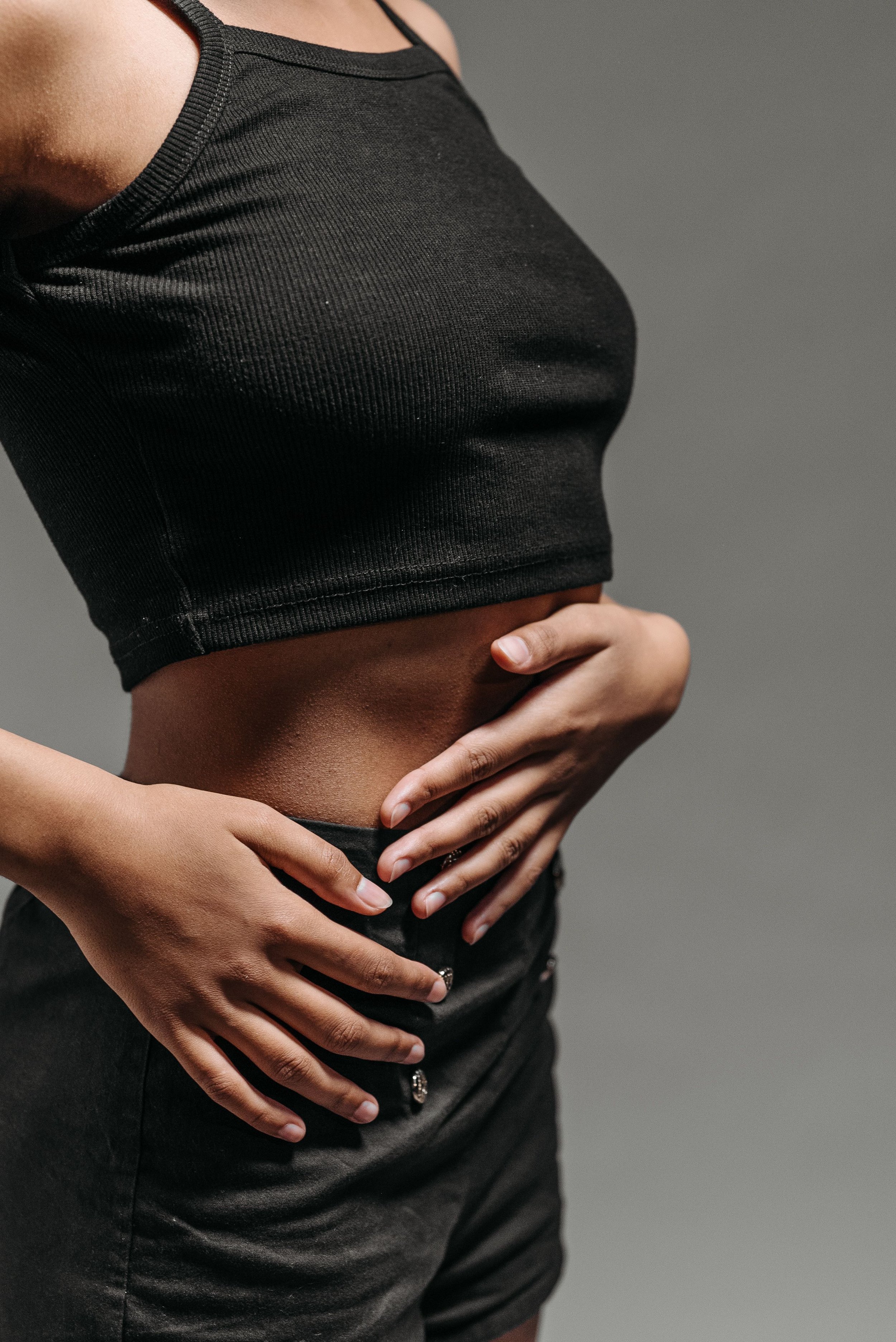 Close-up of a person wearing a black sleeveless top and high-waisted black pants, with their hands resting on their stomach, against a gray background.