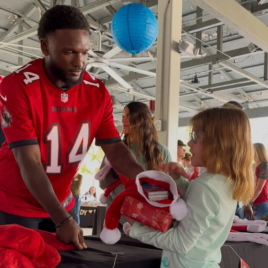A man in a red football jersey is handing a Christmas gift box to a girl with red hair and glasses, under a decorated space with a blue paper lantern hanging from the ceiling.