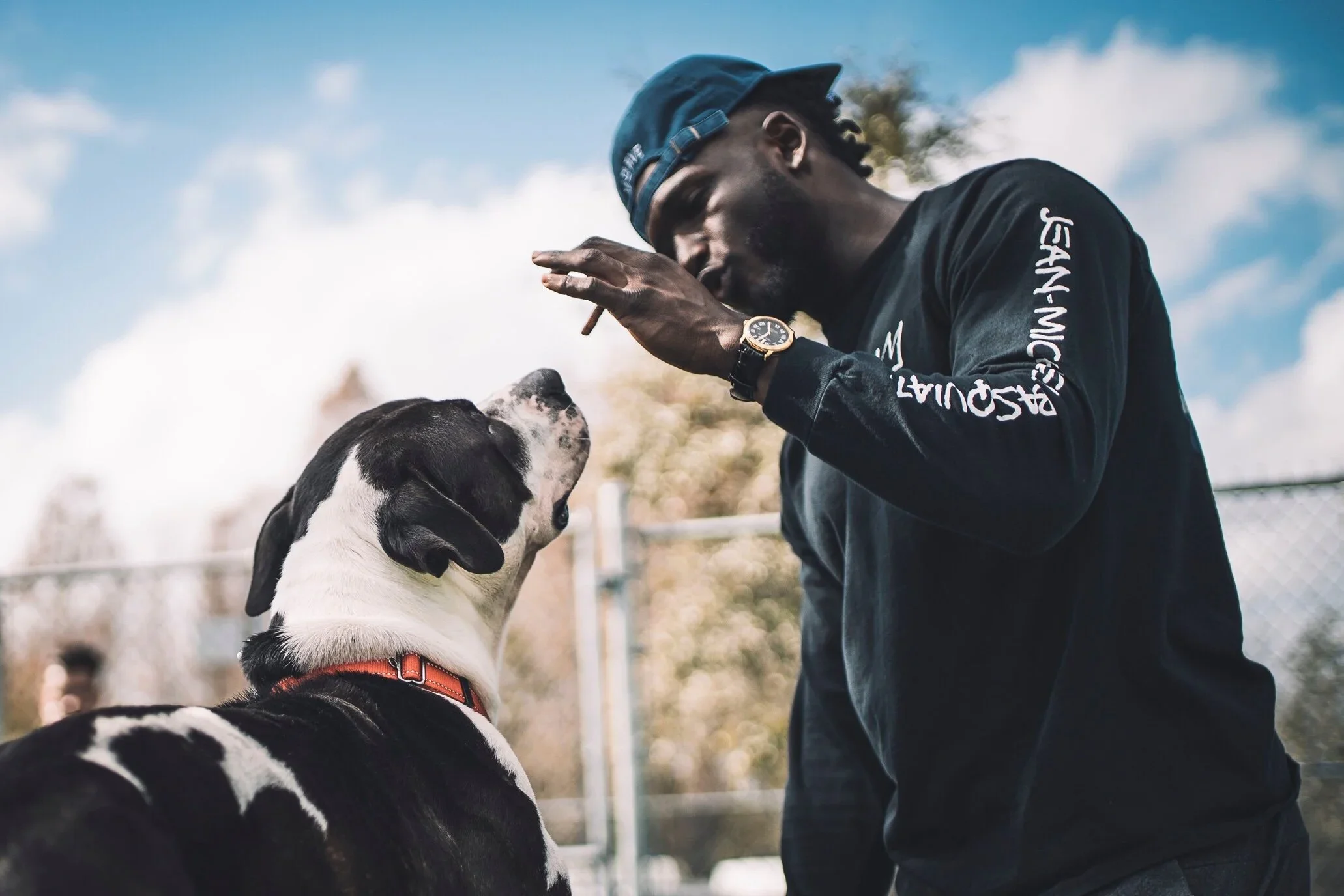 A man wearing a blue cap and black sweatshirt with white text is raising his hand and holding a cigarette, while a black and white dog with a red collar is looking up at him.