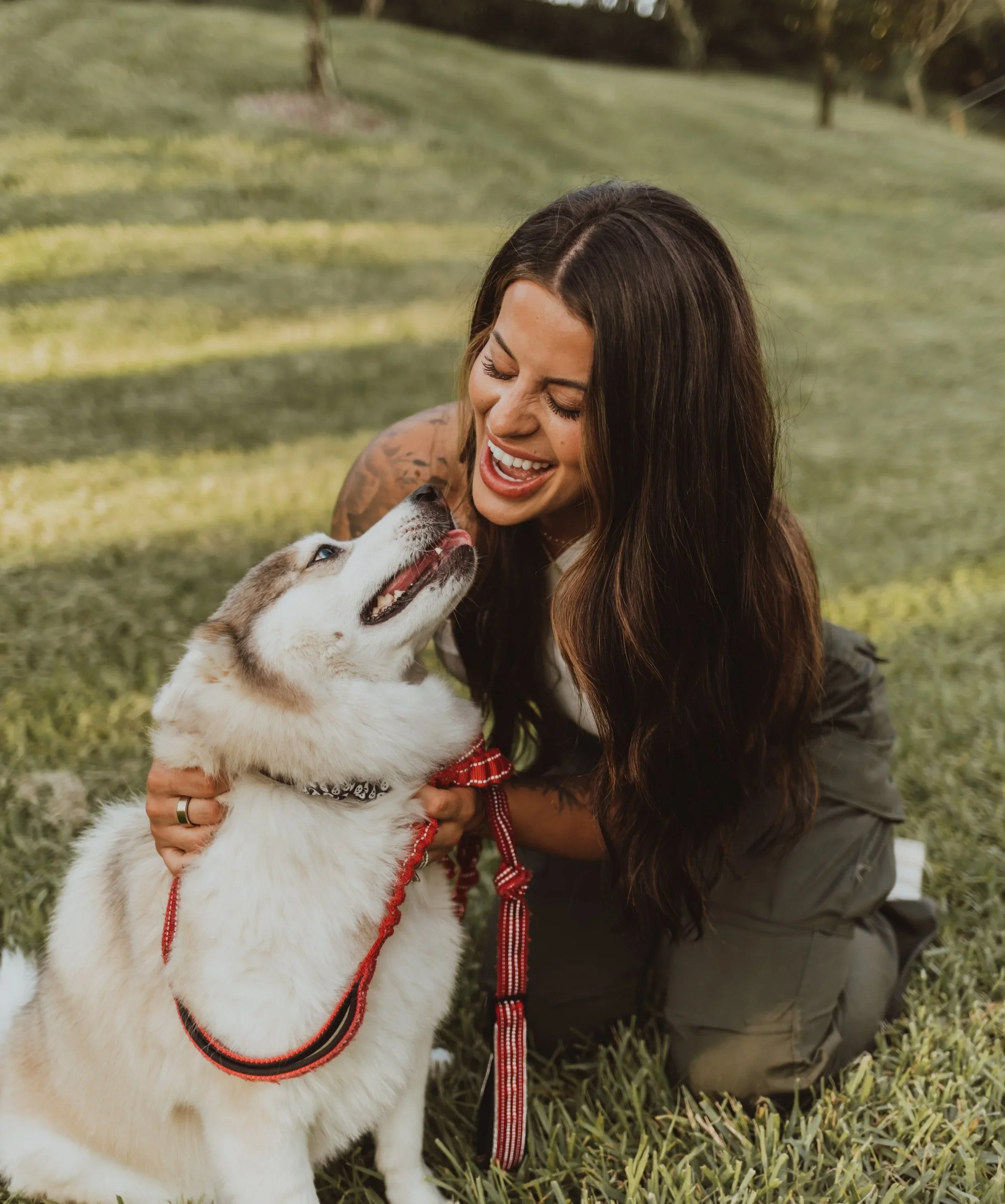 A woman with long dark hair and tattoos smiling while playing with a Siberian Husky puppy outdoors on grass.