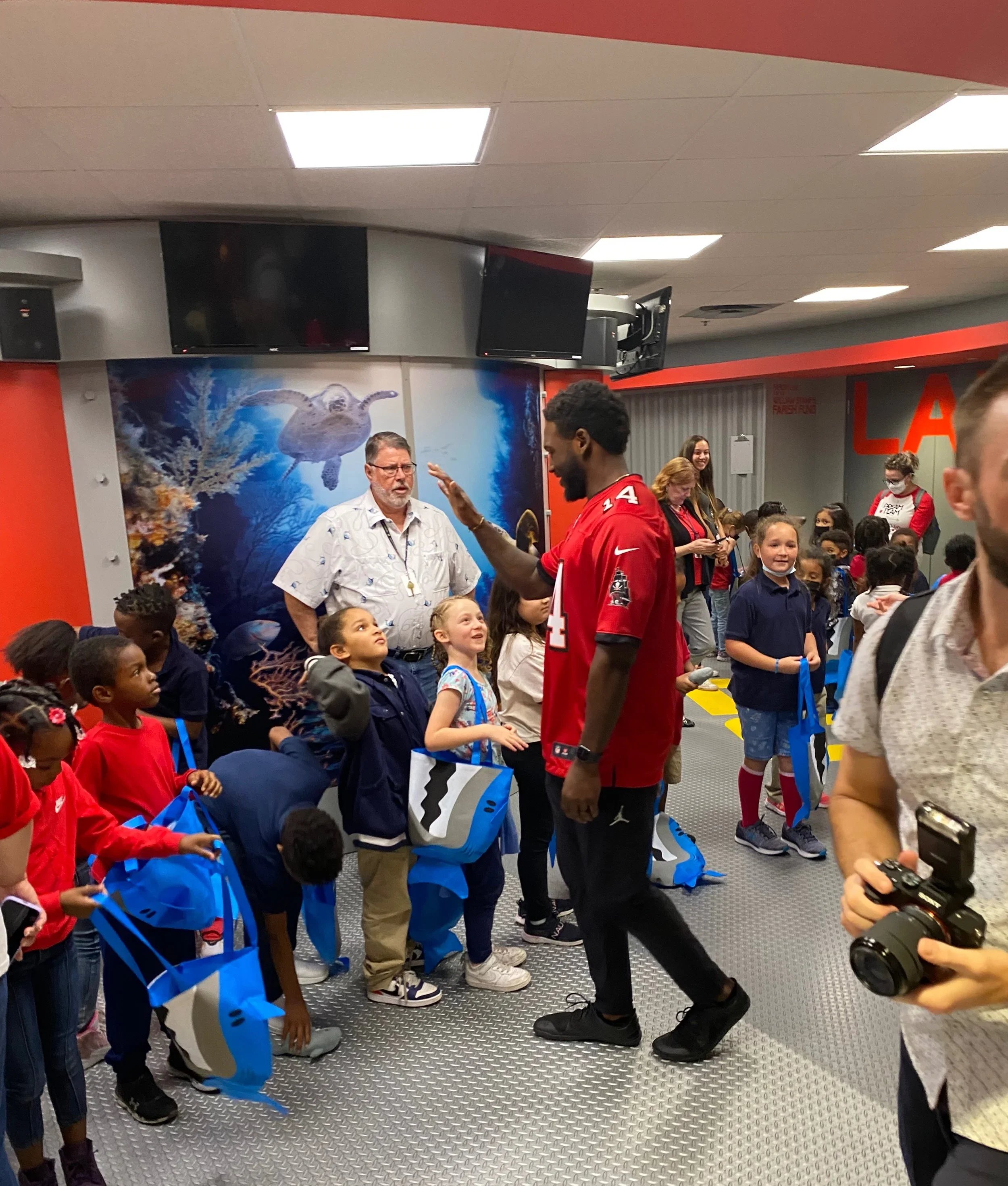 A group of children in a room with a coral reef backdrop, listening to a man in a red sports jersey who is talking to them, with some adults and a photographer also visible.