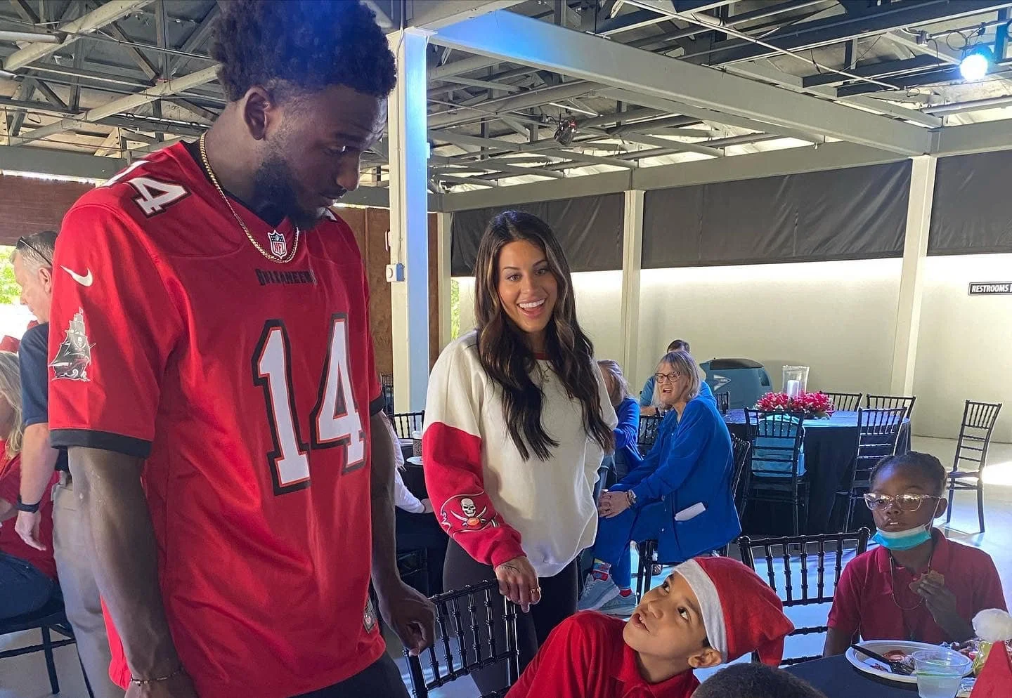 A group of people, including a man in a red football jersey, a woman in a cream and red sweatshirt, and children with festive holiday attire, gathered around a table during a holiday event.