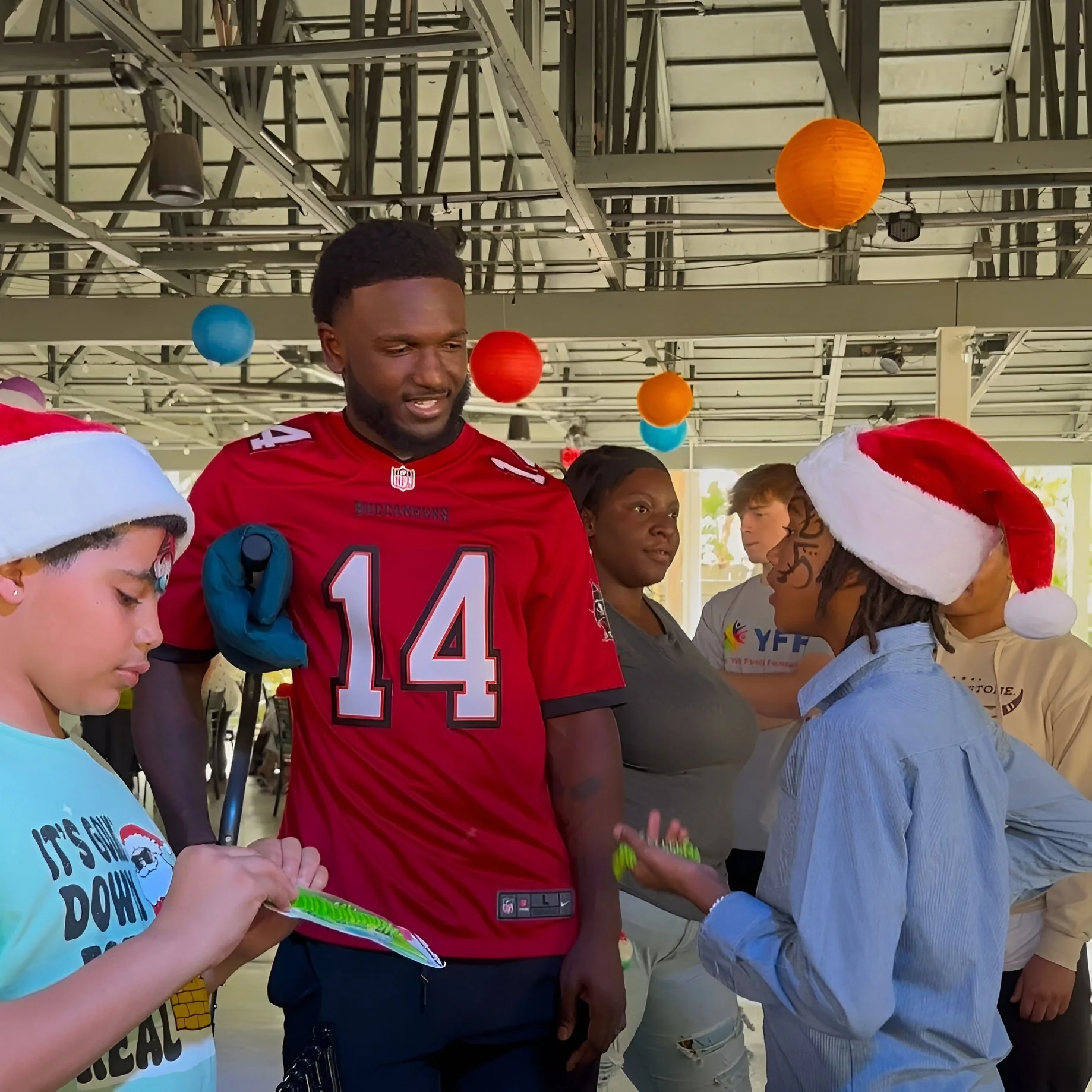 People in a festive setting with colorful hanging lanterns, some individuals wearing Santa hats, and a young man in a sports jersey being engaged in conversation.