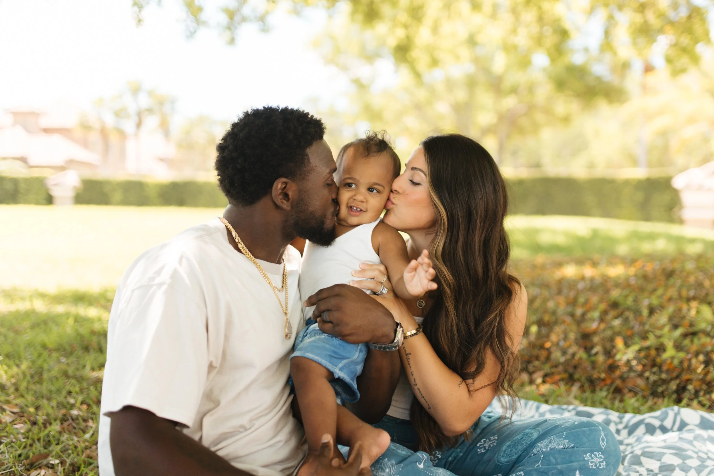 A family of three sitting on a blanket outside, a man and woman kissing their young child on the cheeks with a park and trees in the background.