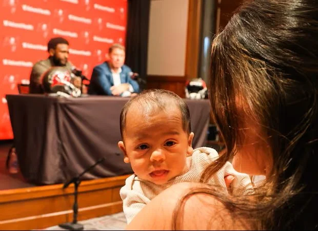 A woman holding a crying baby in front of a panel discussion or press conference with three men sitting at a table on a stage. The backdrop has red branding or logos.