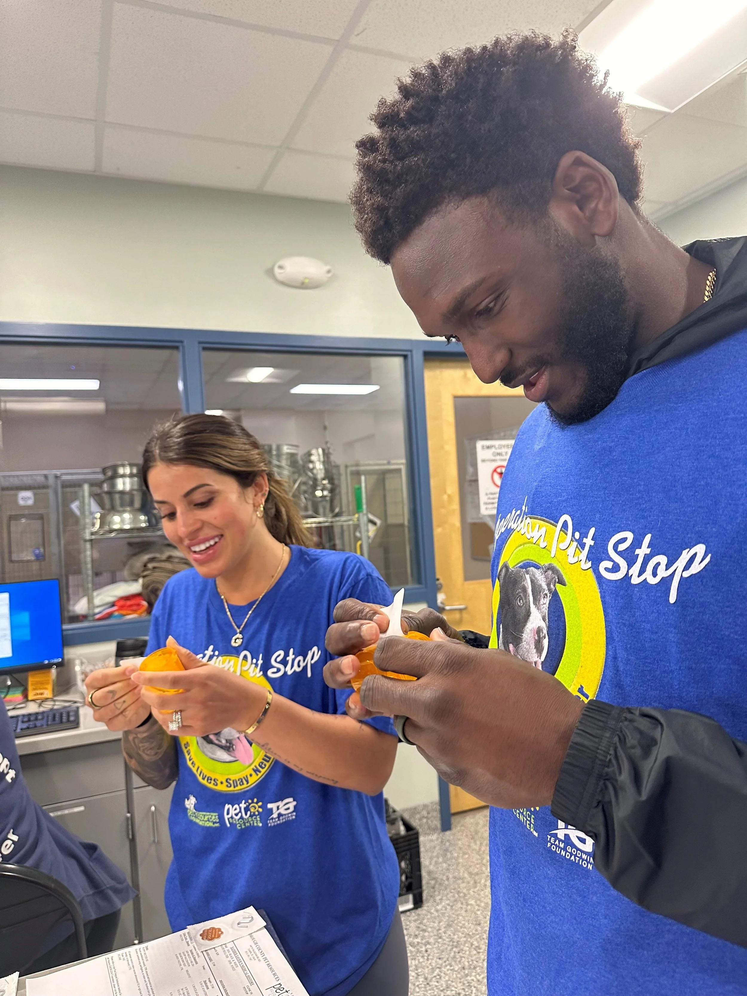 Two smiling volunteers, a woman and a man, wearing blue shirts with 'Animal Pit Stop' logo, are filling orange dog bowls in an animal shelter.