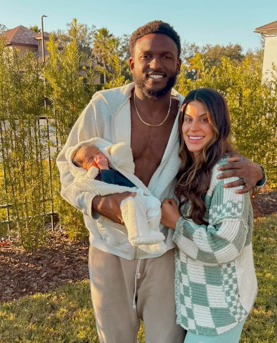 A happy couple holding a newborn baby outside in a garden with green shrubs and trees, during golden hour sunlight.