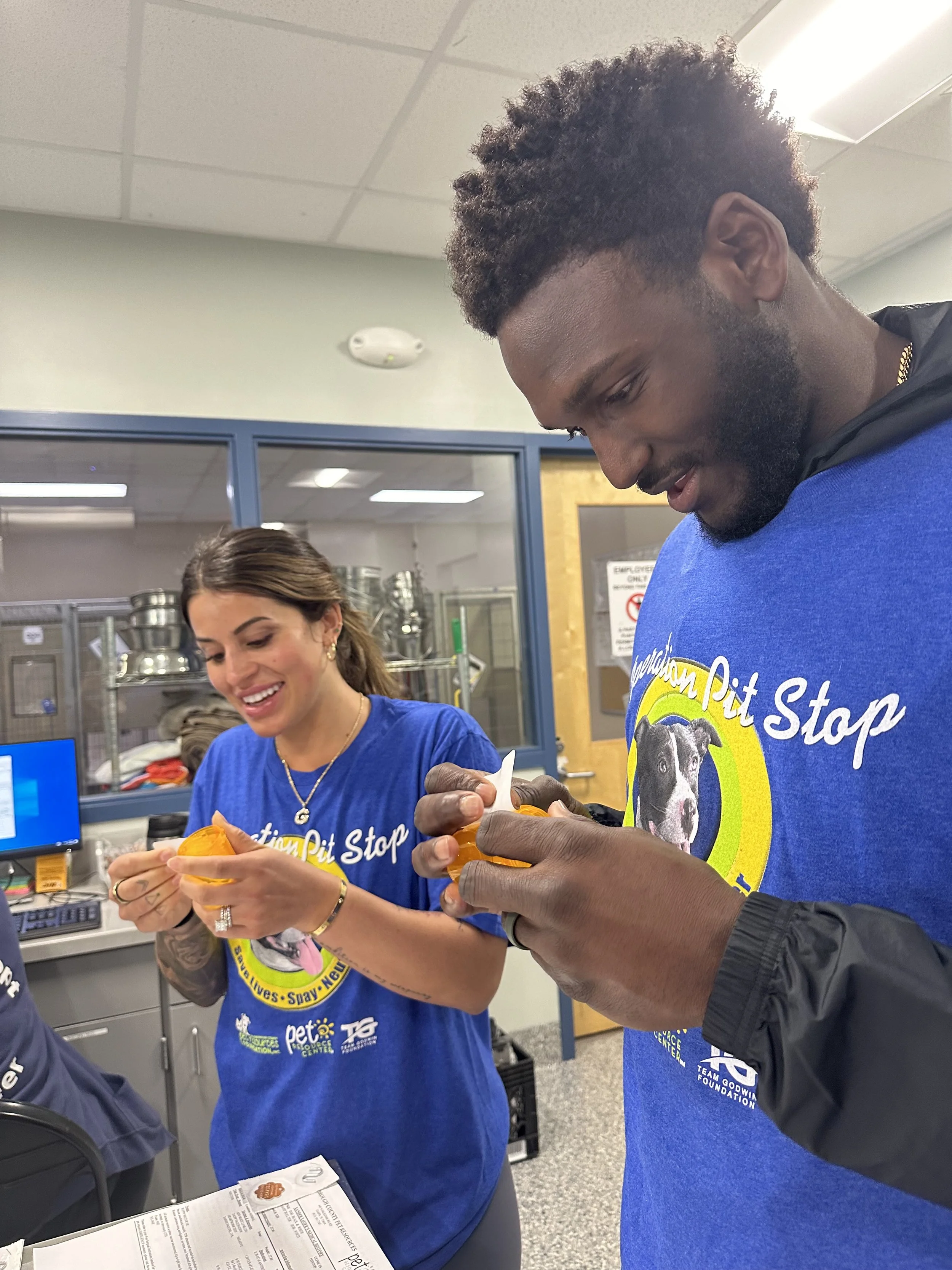 Two volunteers in blue t-shirts labeled 'Animal Pit Stop' are decorating cupcakes, smiling indoors with a window and kitchen area in the background.