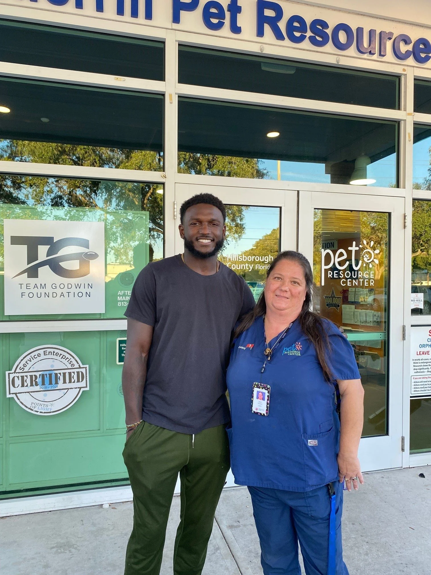 A man and a woman stand outside the entrance to a pet resource center, smiling for the camera. The woman is wearing a blue uniform with an ID badge, and the man is dressed casually in a black T-shirt and green pants.