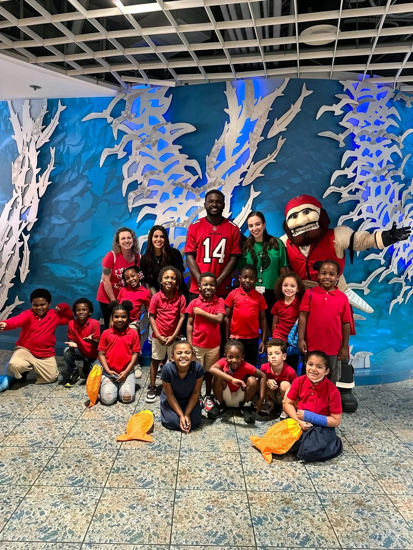 Group of children and adults posing in front of a blue underwater-themed mural with a mascot dressed as a pirate, some children are holding inflatable fish.