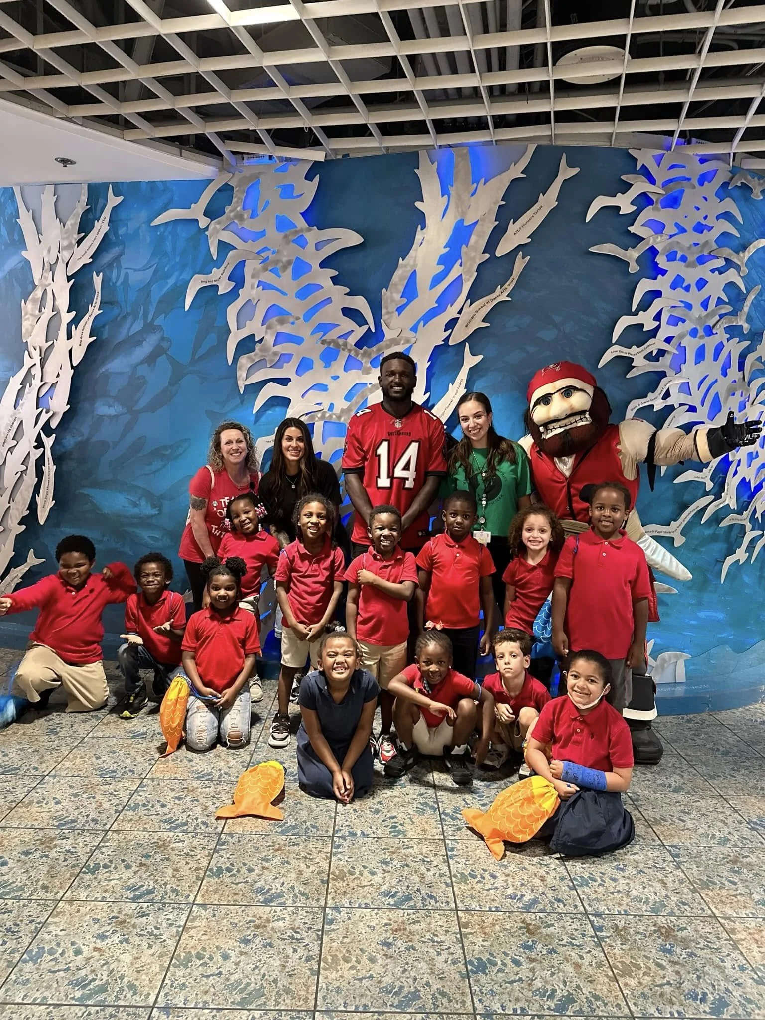 A group of children and adults posing in front of a blue and white decorative backdrop with a mascot character. The children are wearing red shirts and some are holding orange fish-shaped objects. The adults include a woman with curly hair, a man in 