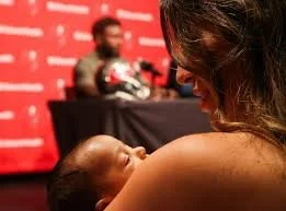 A woman holding a newborn baby in a room with a red wall and a man in the background.