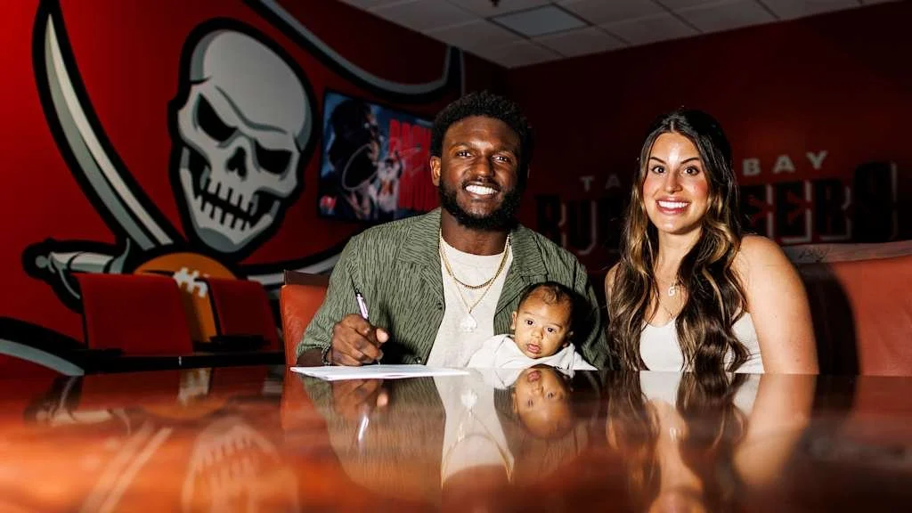 A family of three sitting at a table in a Tampa Bay Buccaneers restaurant, with a large Buccaneers logo on the wall behind them.