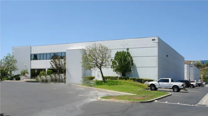 A large, white commercial or industrial building with parking lot and a few trees in front, under a clear blue sky.