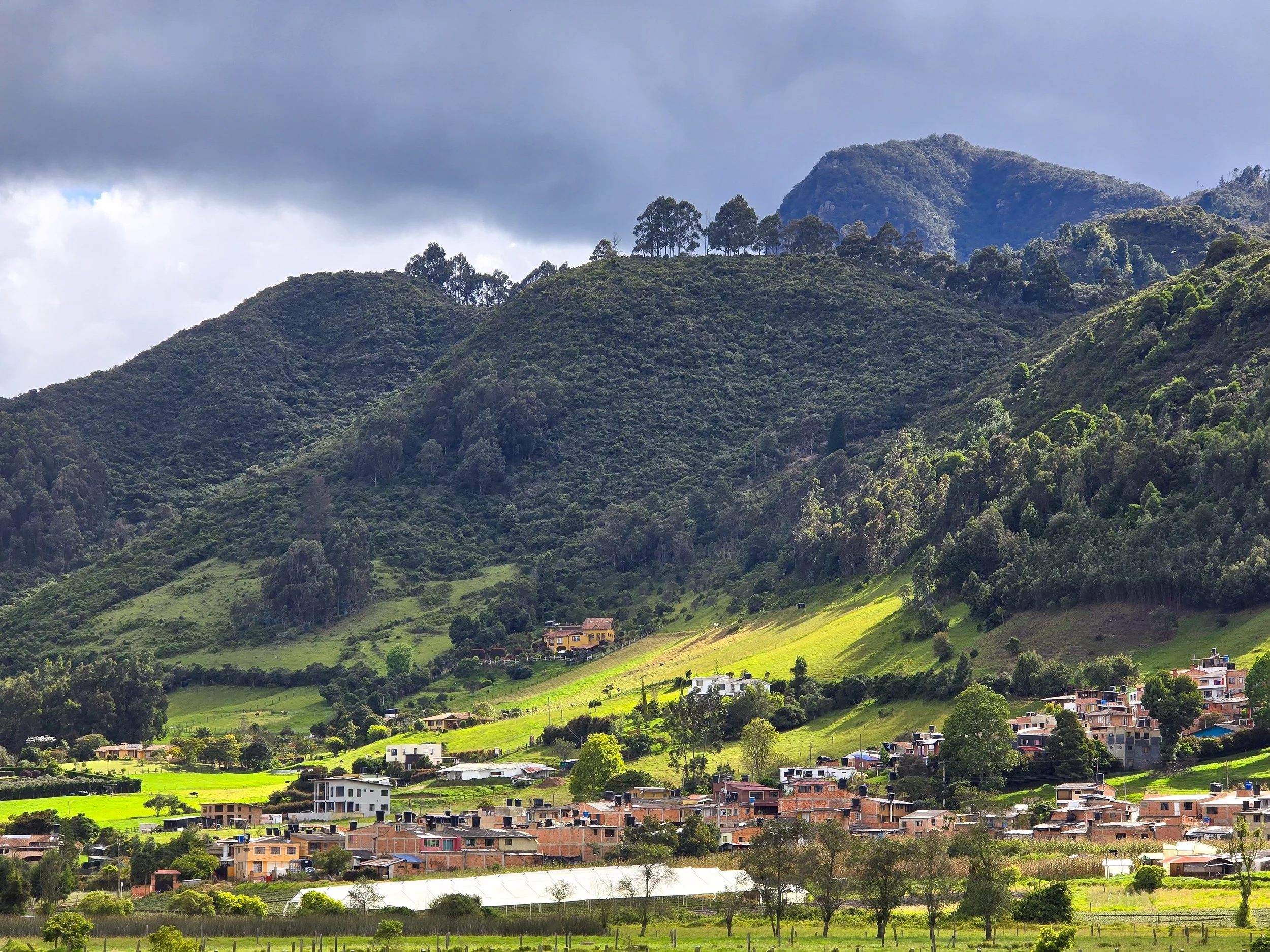 Green hillside village surrounded by lush forested mountains and partly cloudy sky.