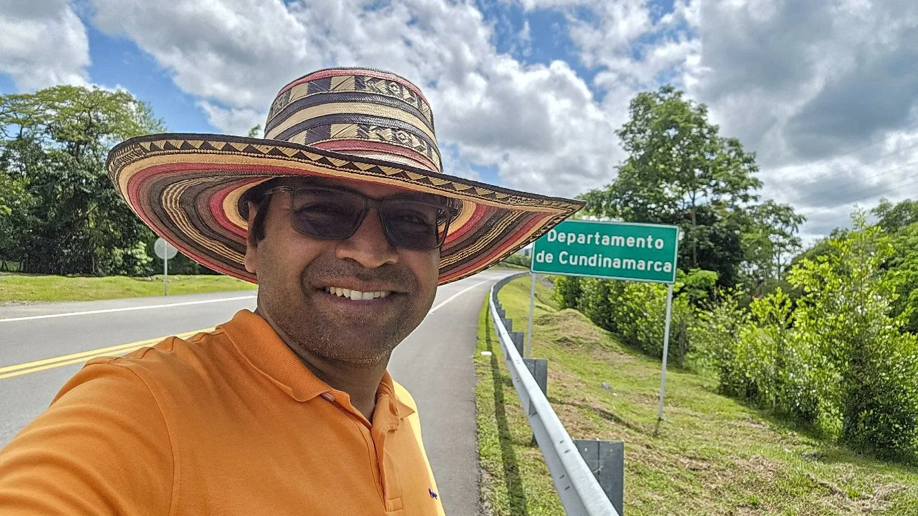 A man smiling, wearing a large colorful hat, sunglasses, and an orange shirt, standing on the side of a road with a green sign that reads 'Departamento de Cundinamarca,' surrounded by trees and cloudy sky.