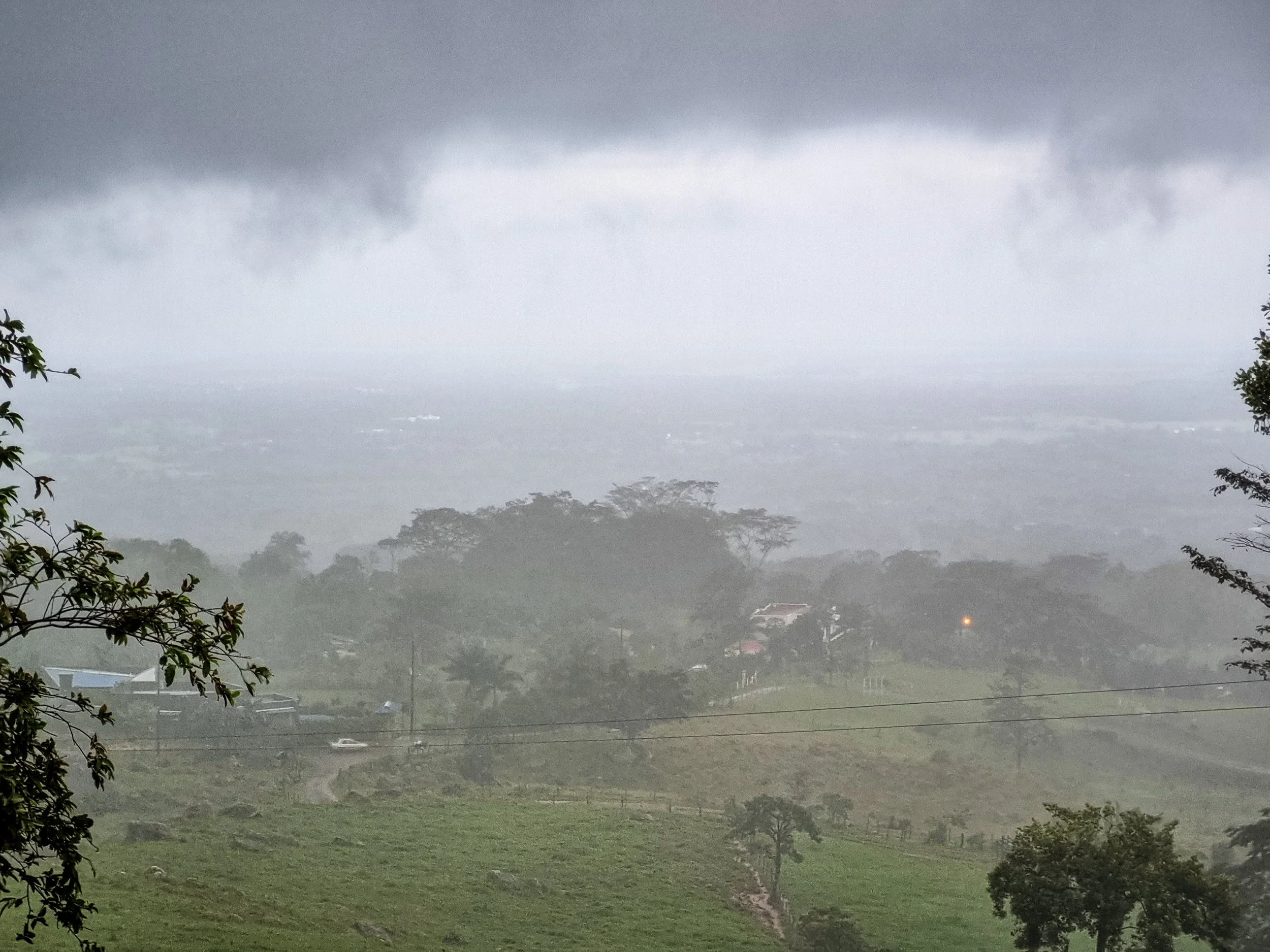 A scenic view of a lush green countryside with trees, fields, and a few houses. Dark storm clouds hang overhead, creating an intense and moody atmosphere.