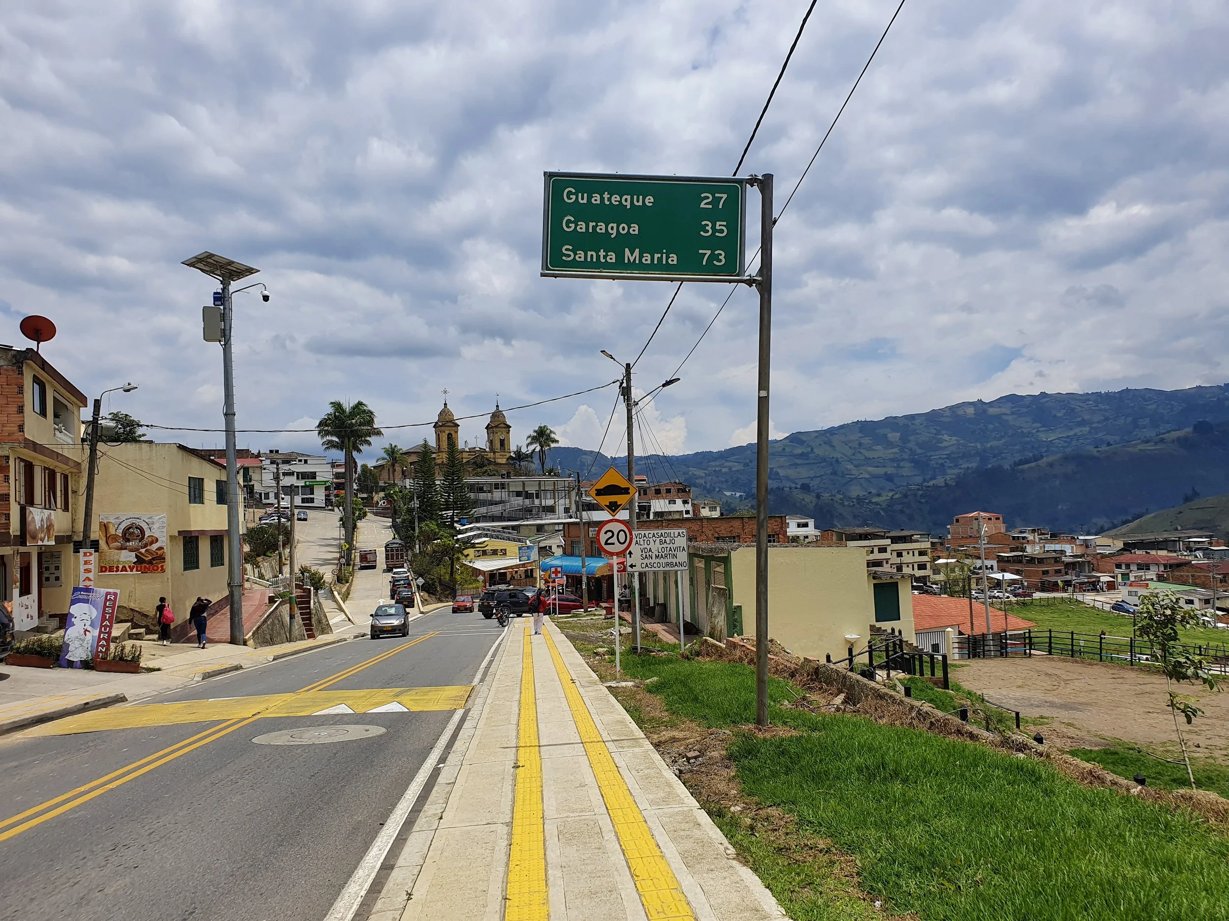 Street view with a green directional sign indicating distances to Guateque, Garagoa, and Santa Maria, in a hilly town with buildings, palm trees, and mountains in the background.
