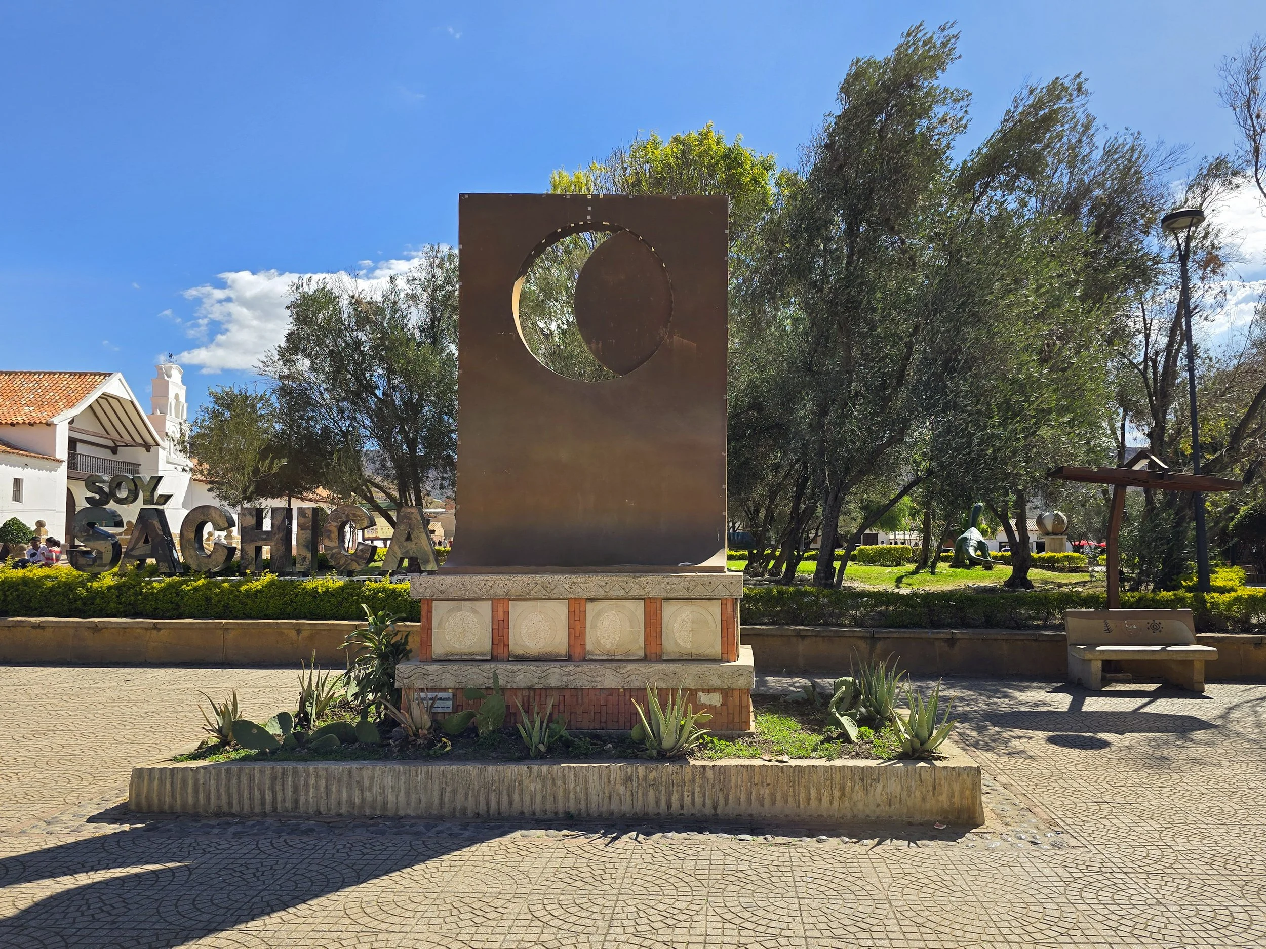 Public park with a modern sculpture featuring a large rectangular metal panel with a circular cutout, surrounded by trees, a bench, and decorative plants. In the background, there are buildings with signs, including one that says "SOY SACHICA." The sky is blue with some clouds.
