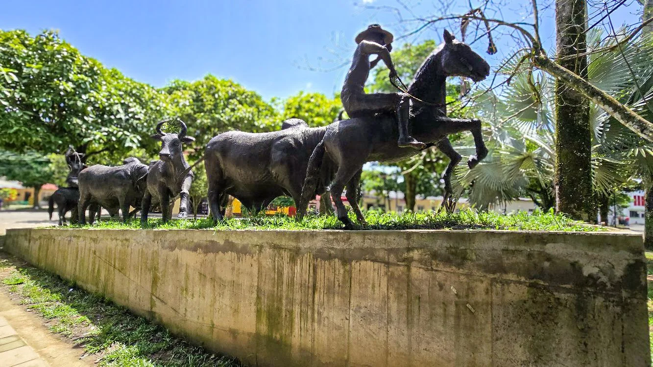 A bronze sculpture of a herd of cattle and a mounted cowboy in a park with green trees and a blue sky in the background.