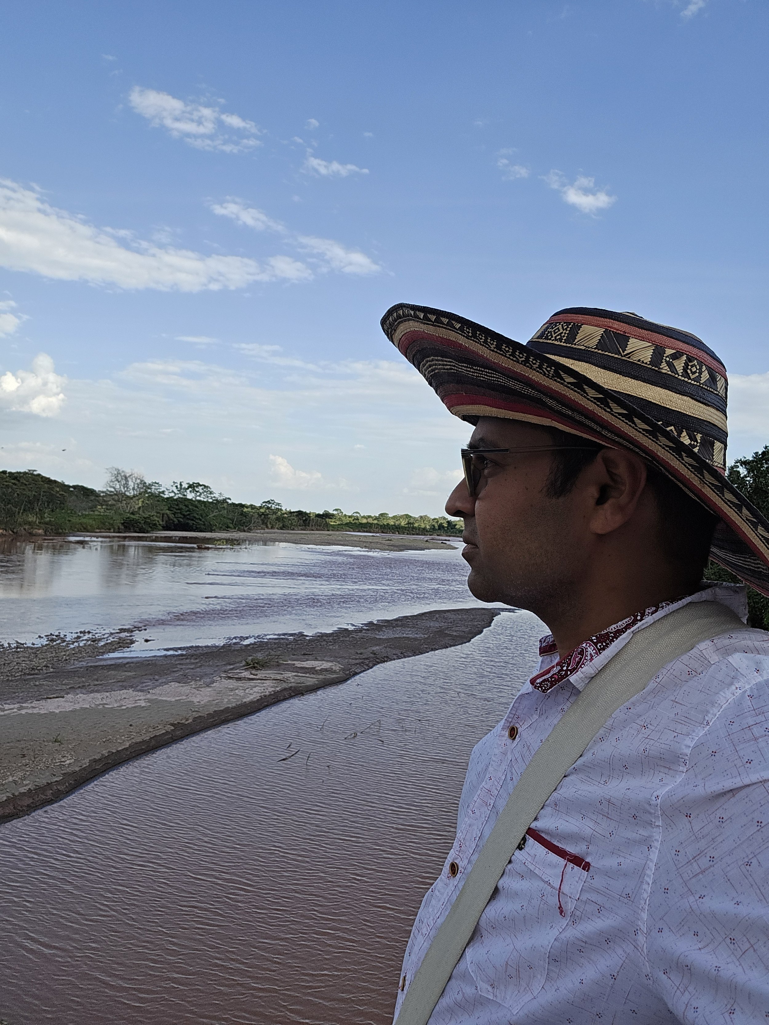A man with glasses and a wide-brimmed, colorful, patterned hat stands near a river, looking into the distance during daytime.