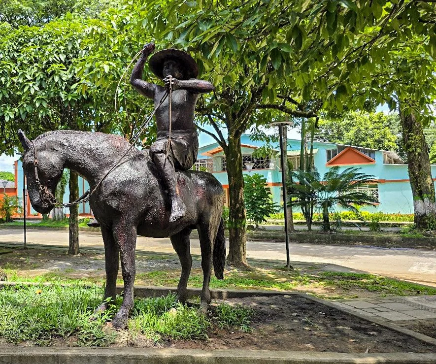 Statue of a cowboy riding a horse, located outdoors with trees and a building in the background.