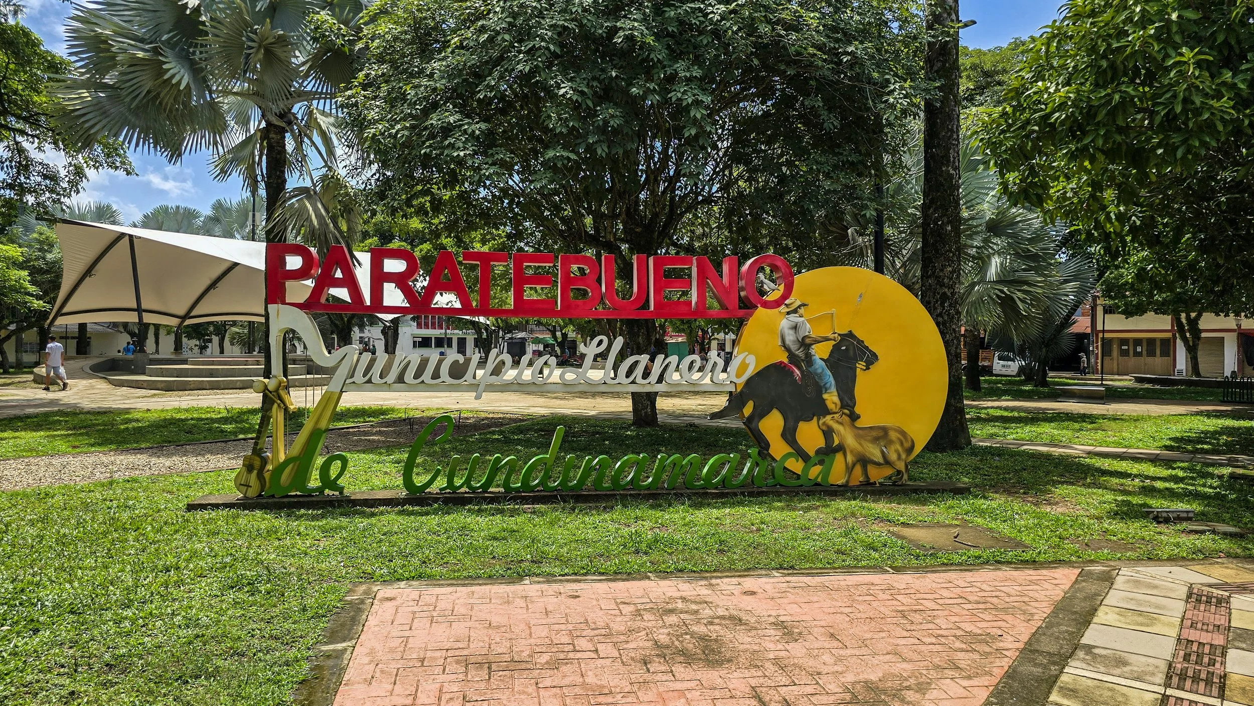 Sign for Parate Bueno, Municipalidad Lano de Cundinamarca, with a painted figure of a man on horseback, a dog, a yellow circle, green grass, trees, and a paved walkway.