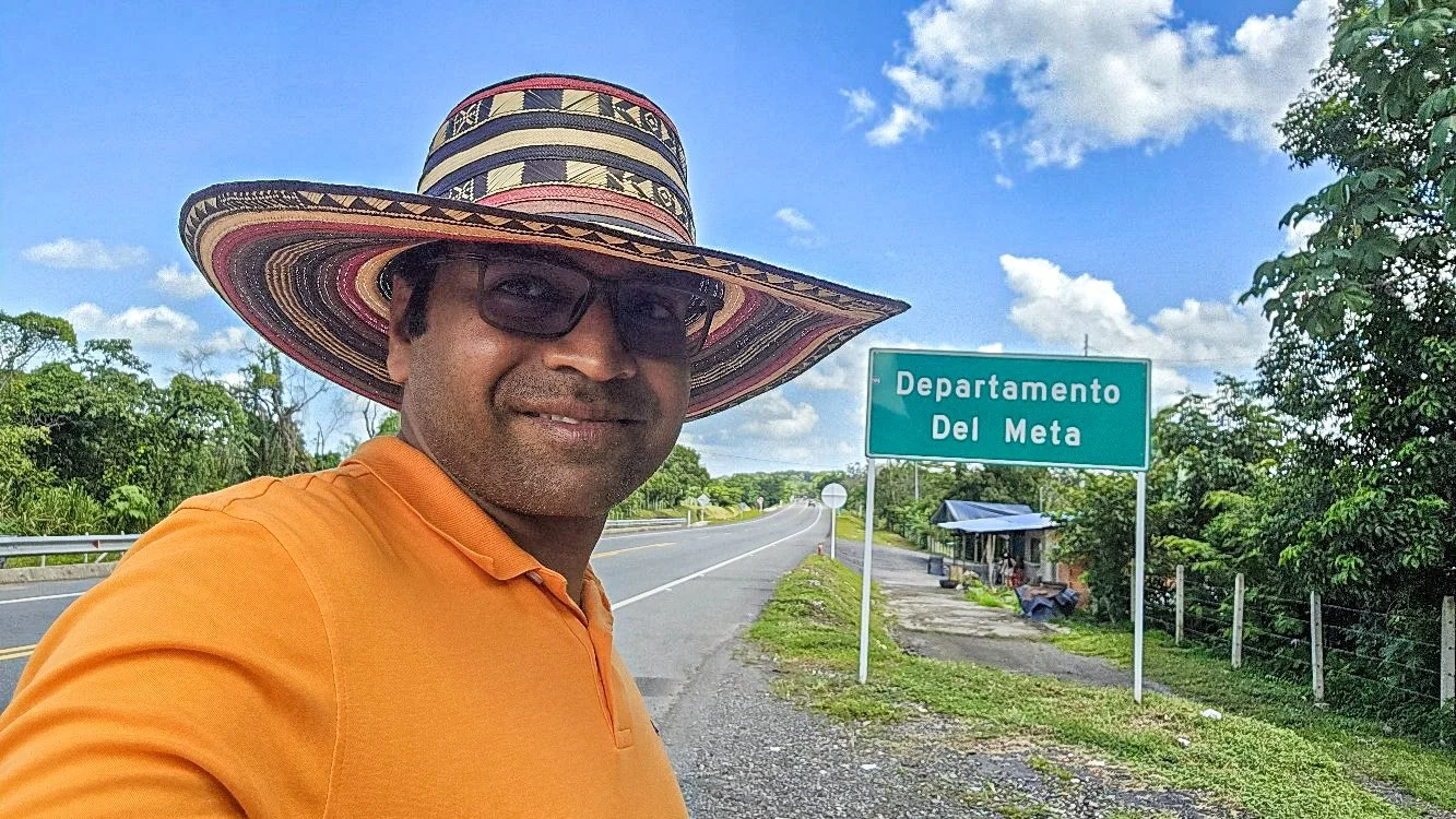 A man wearing a colorful woven hat and sunglasses takes a selfie on the side of a rural road with a sign that reads 'Departamento Del Meta' in the background, lush green trees, and a partly cloudy sky.