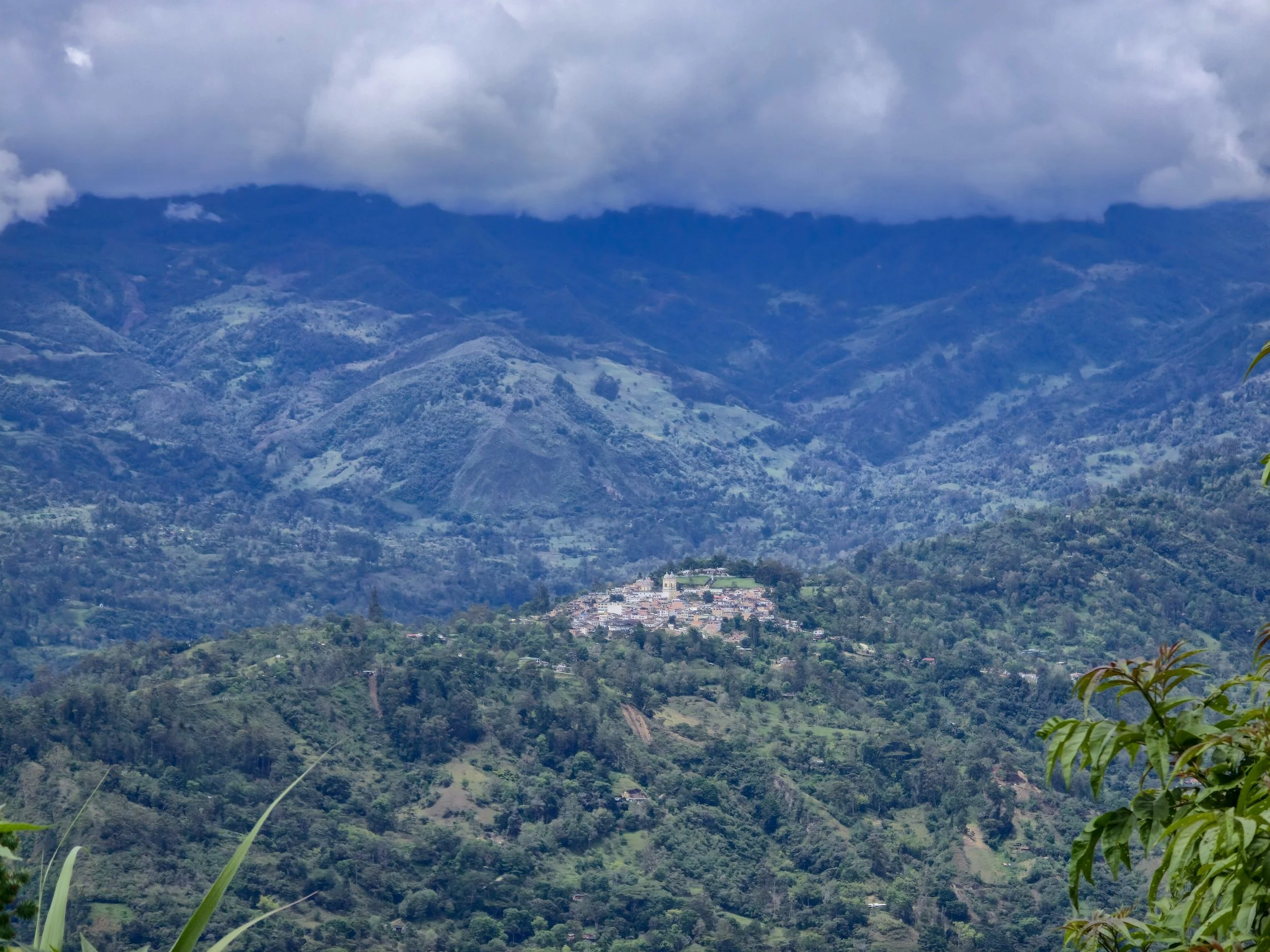 A mountain landscape with clouds above, and a small village nestled on a hillside covered in lush green vegetation.