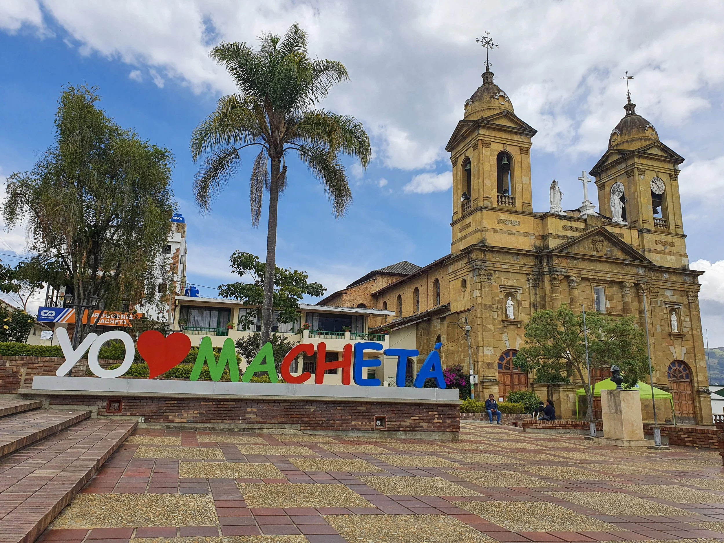 Colorful sign that says "Yo corazón Machetá" in a town square with a historic church, palm trees, and a few people sitting and walking around, bright sky with some clouds.