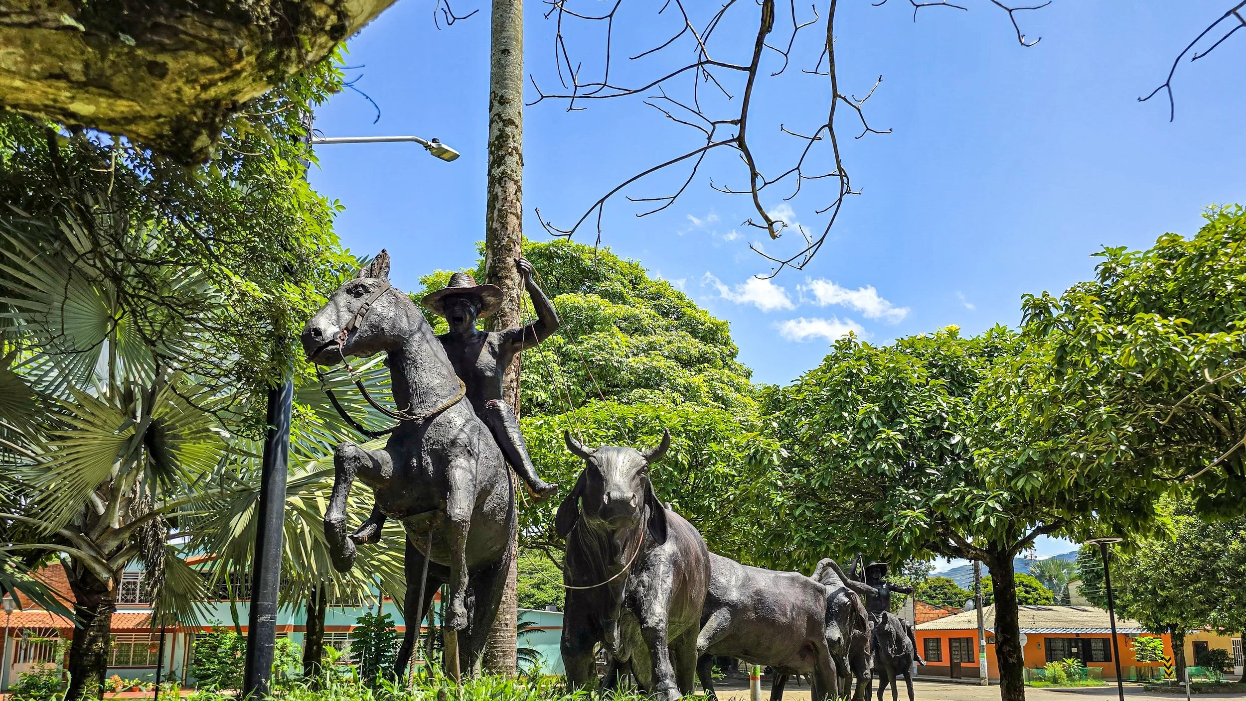 Statues of a cowboy riding a horse leading a herd of cattle in a park with trees and a building in the background.