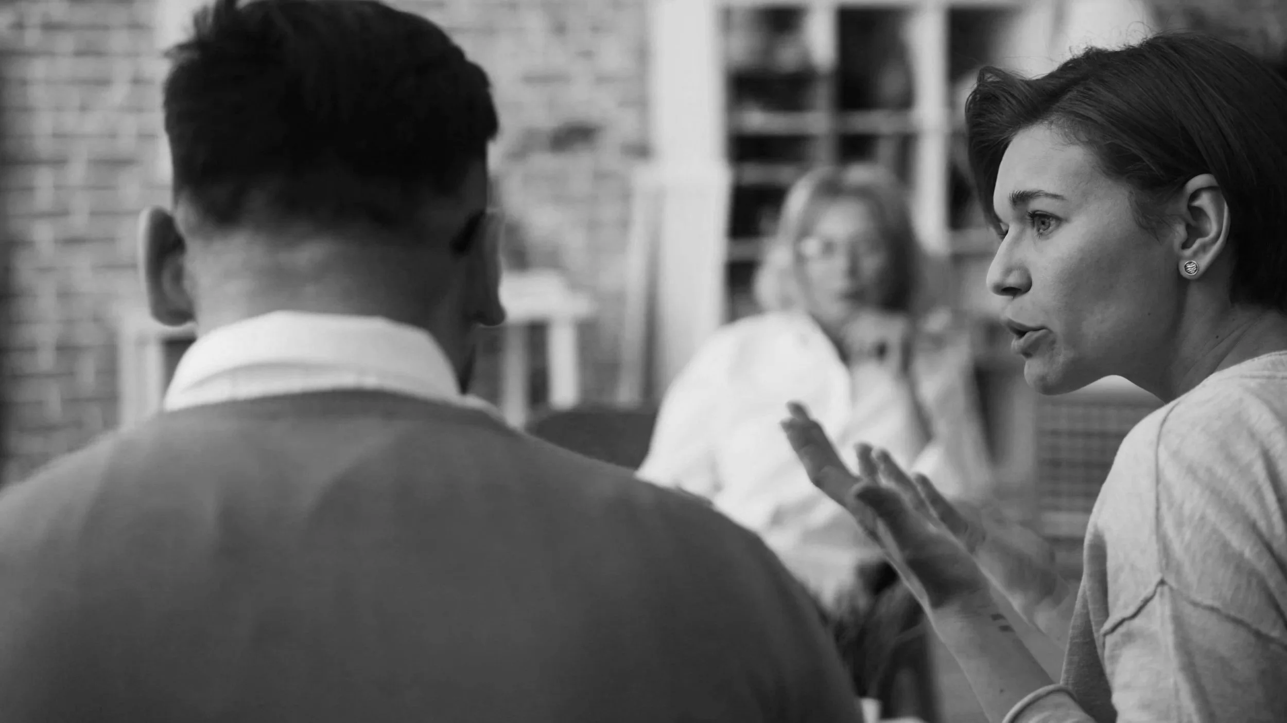 Black and white image of 3 people on a meeting and a woman speaking to a man with her hands and an angry face. The third person is watching.