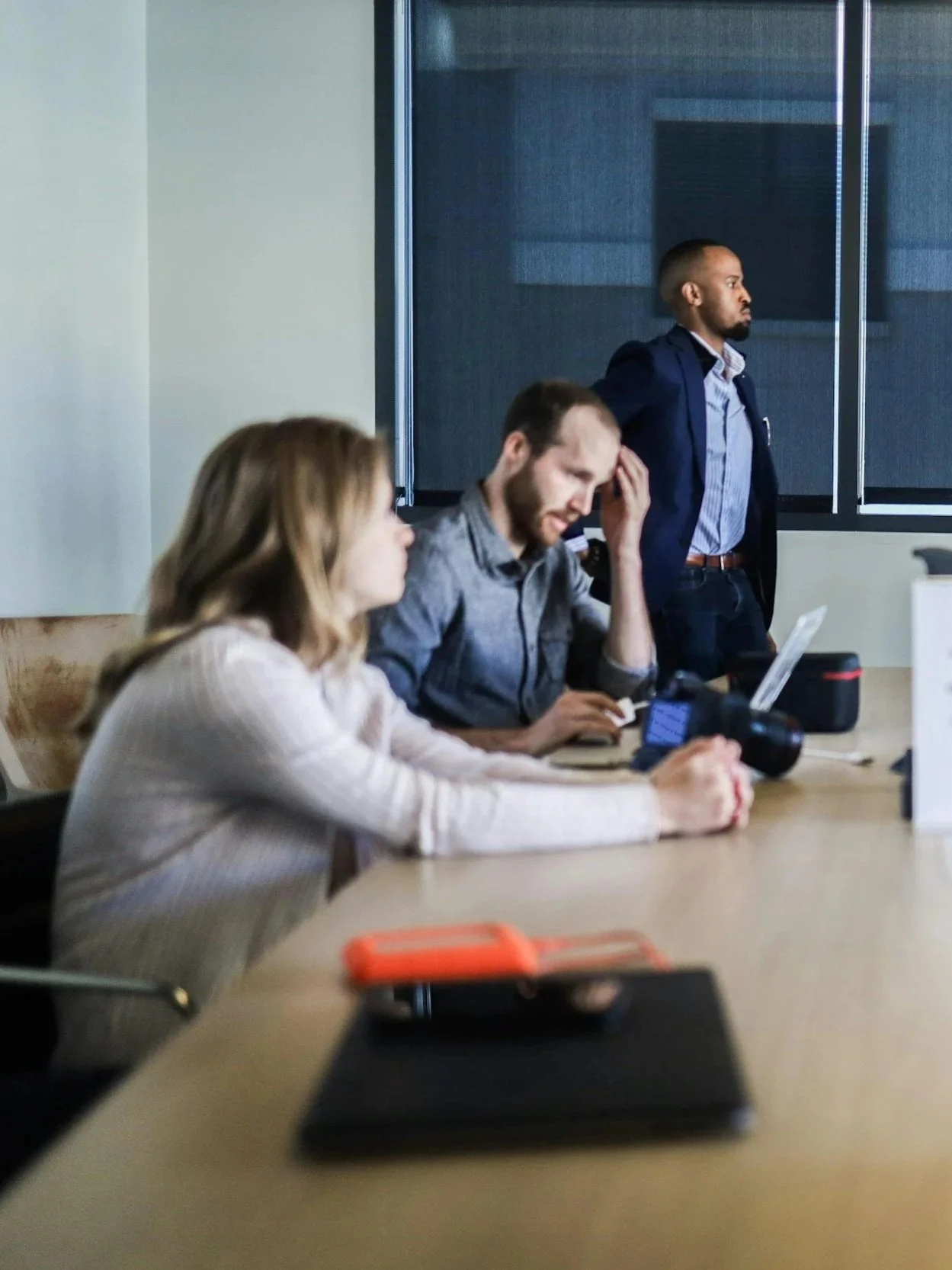 Corporate team members in a business meeting showing signs of conflict — one standing with a scowl, one seated with head in hand, another disengaged and looking away.