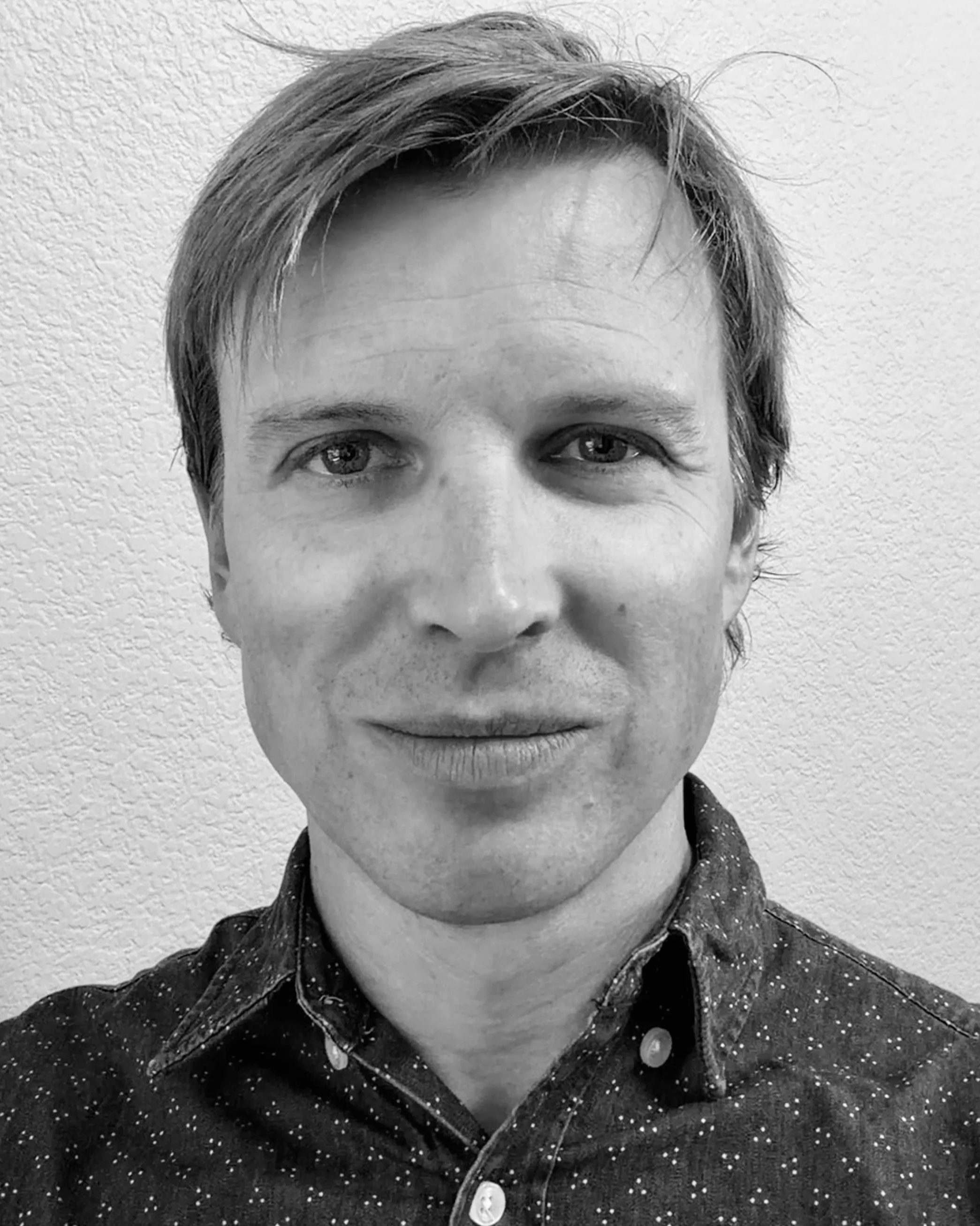 Close-up black and white photo of a person's face, showing the forehead, eyes, nose, and mouth. The person has short, slightly tousled hair and is wearing a collared, polka-dotted shirt.