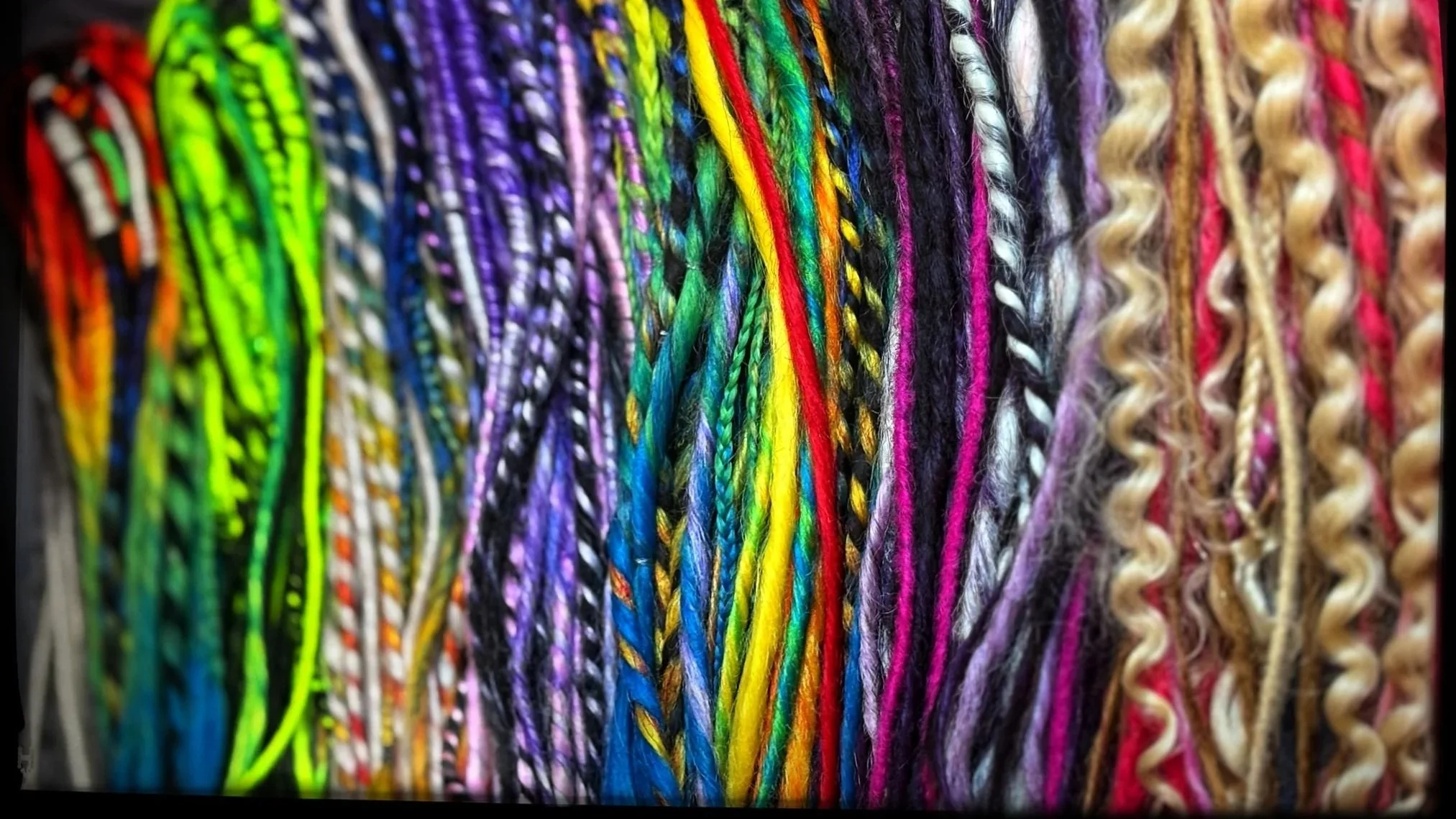 Close-up of various colorful braids and Dreads arranged vertically.