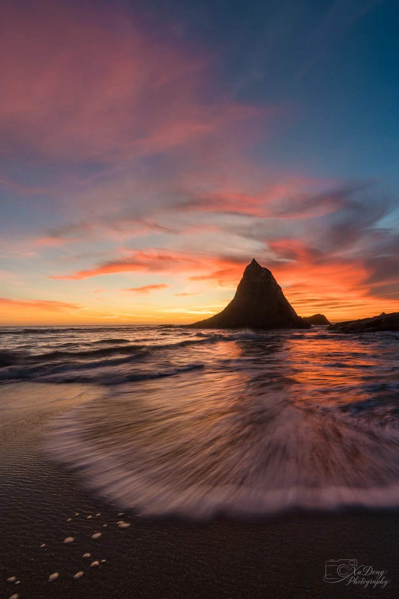 Sunset photography of a coastal spire silhouette with motion-blurred waves washing over the shore