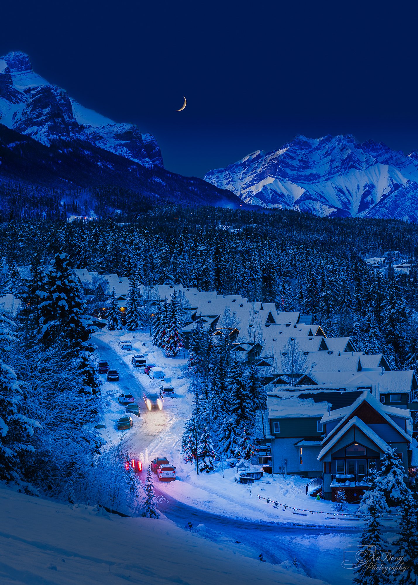 A peaceful winter nightscape of a small mountain village nestled in snow, glowing warmly under the soft, cool light of a crescent moon and deep blue mountain shadows.